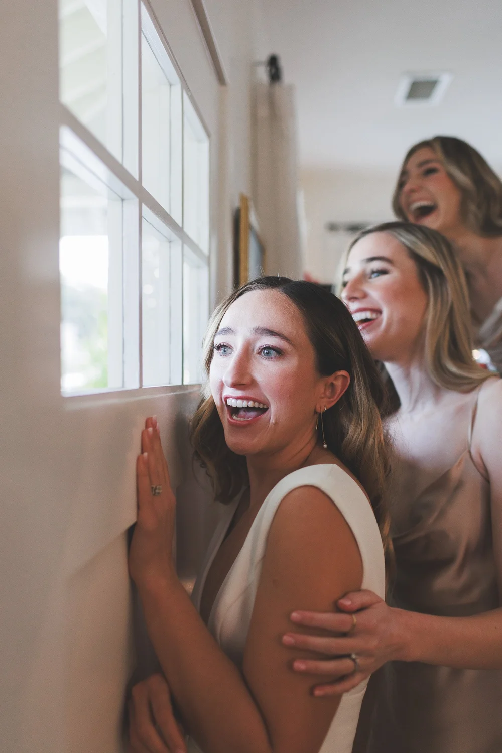 Bridesmaids peering through a window with hands pressed to the glass, laughing and emotional