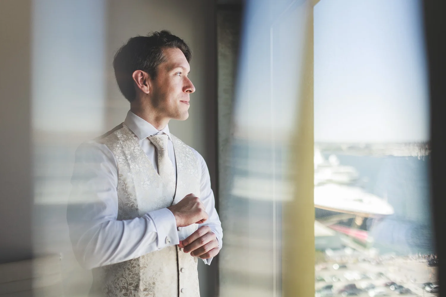 Groom fastening his cuff at a window overlooking a harbour, wearing a brocade waistcoat and pale tie