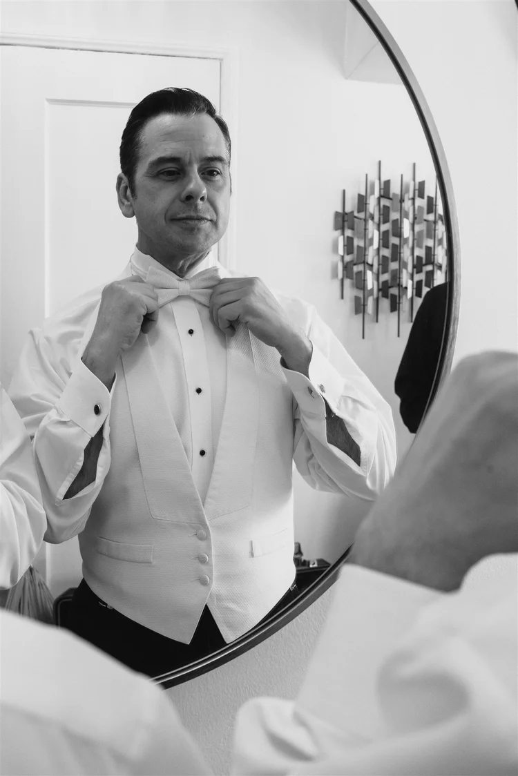 Black-and-white photo of a groom in white tie adjusting his bow tie in a round mirror before James and Beau's wedding
