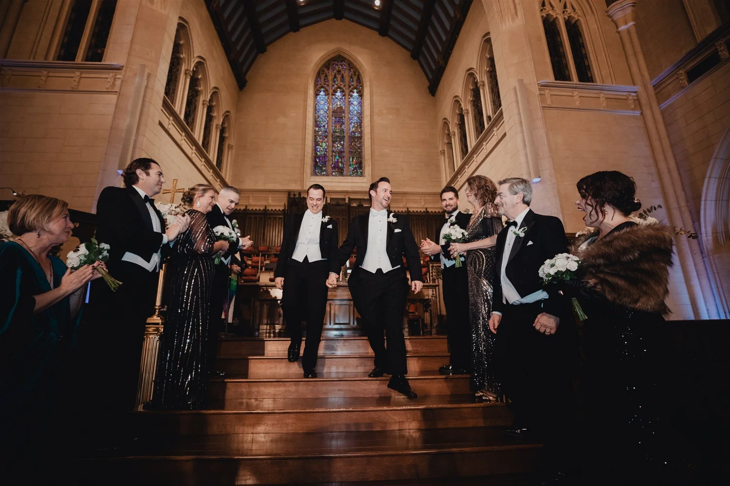 Two grooms in white tie walking down the chancel steps together as their wedding party applauds