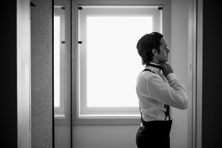 Black-and-white photo of a groom fastening his collar by a tall window before Dylan and Karina's wedding at the Oviatt Penthouse