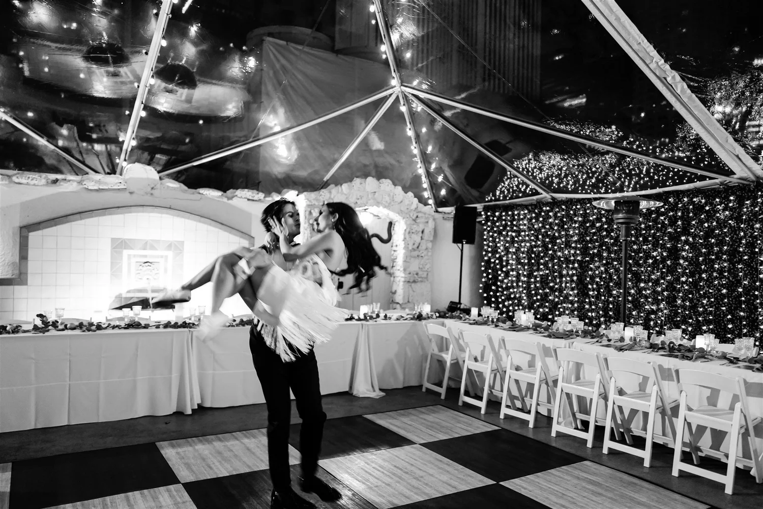 Black and white frame of the groom carrying the bride into a decorated reception tent