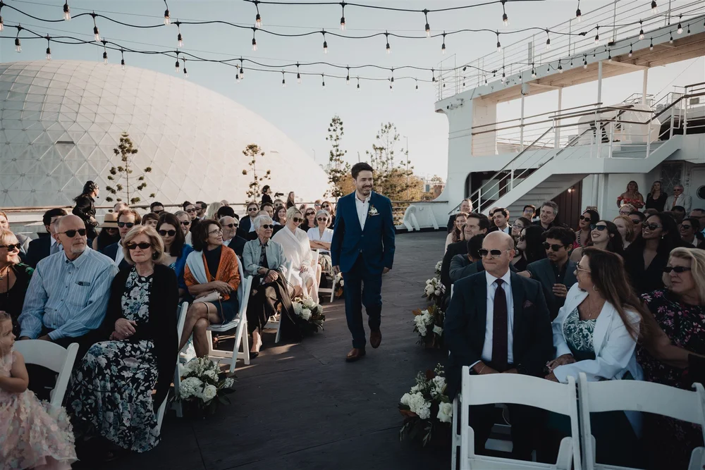 Groom walking the aisle at a deck ceremony aboard the Queen Mary under string lights
