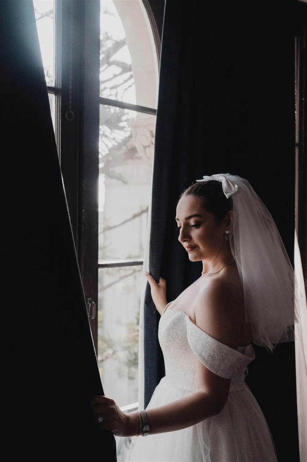 A bride in her veil at a window, holding back the curtain in soft daylight