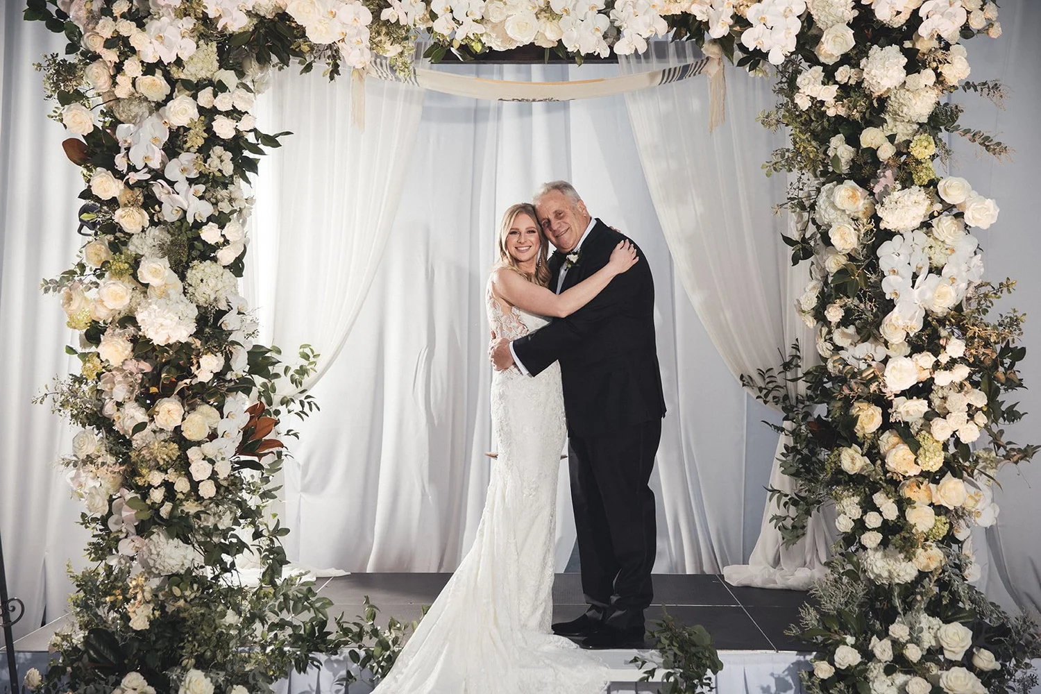 Bride hugging her father beneath a white floral arch at NOOR in Pasadena