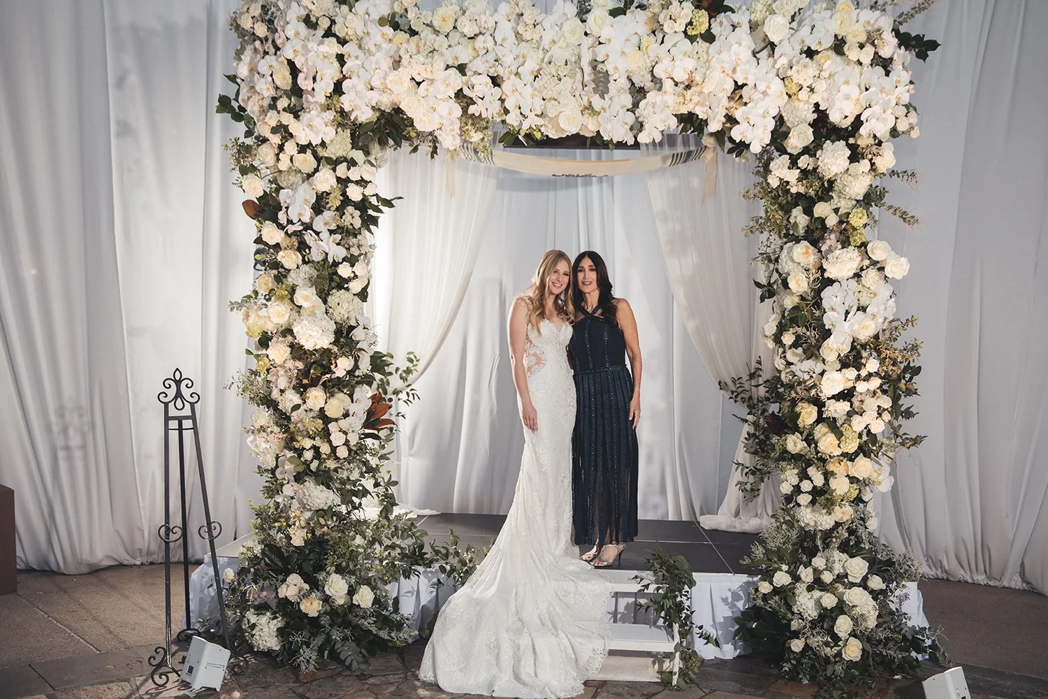 Bride standing with her mother beneath a white floral arch at NOOR in Pasadena