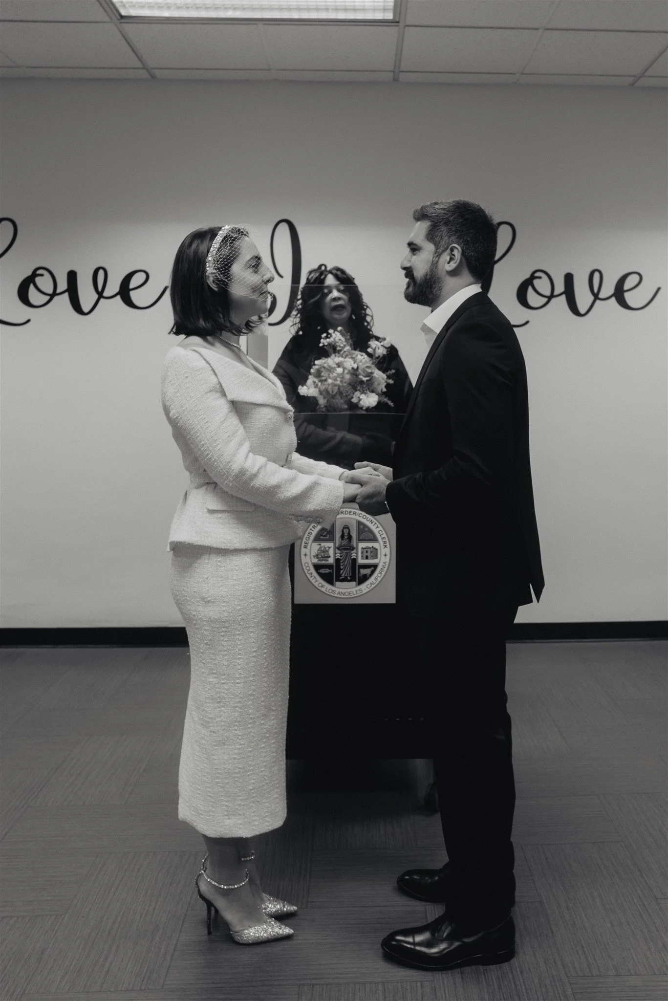 Black and white frame of a couple holding hands during their courthouse ceremony, the word love lettered on the wall behind them
