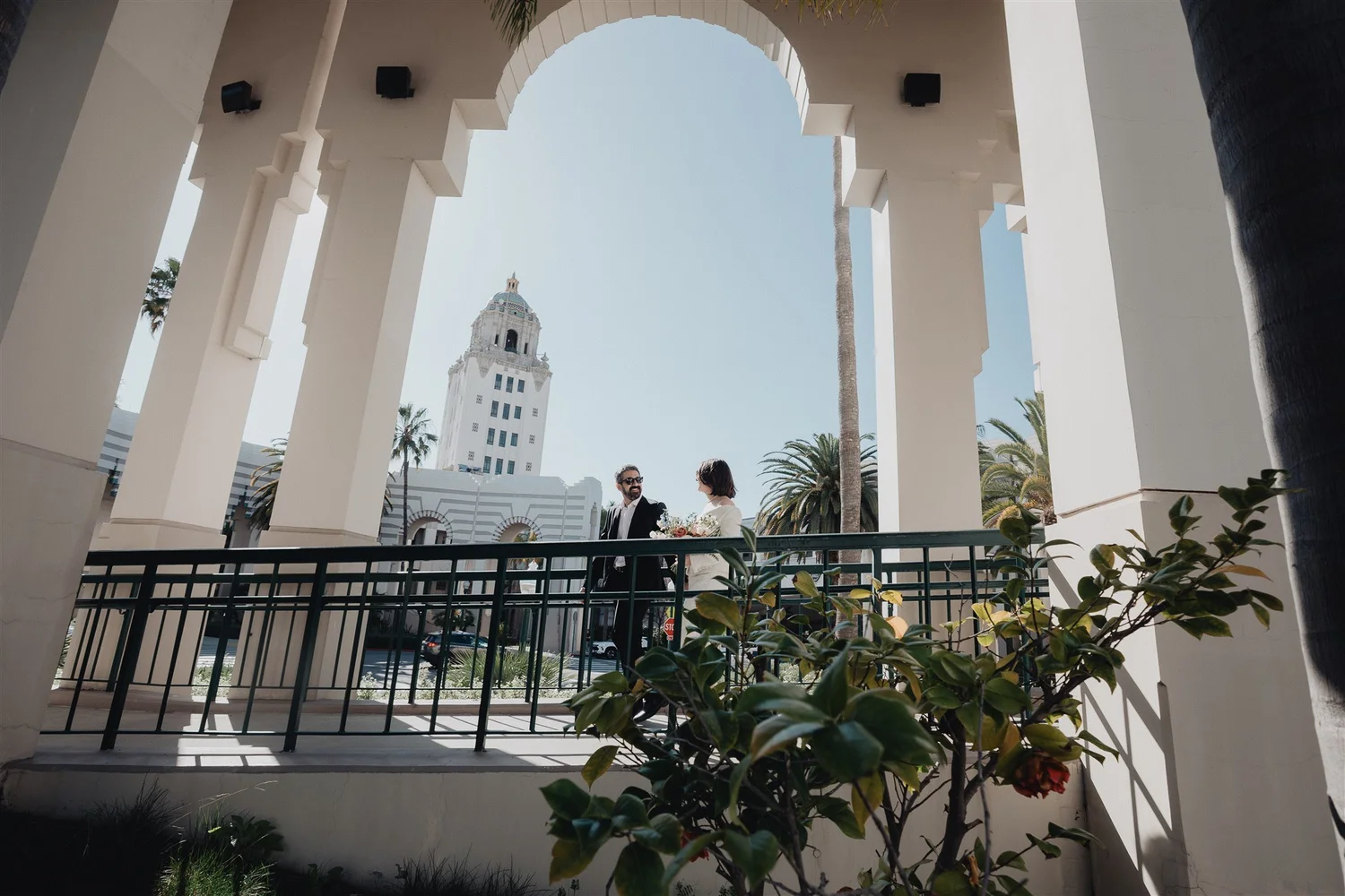 Bride and groom framed by an arched loggia with the Beverly Hills City Hall tower behind them after their Los Angeles courthouse wedding