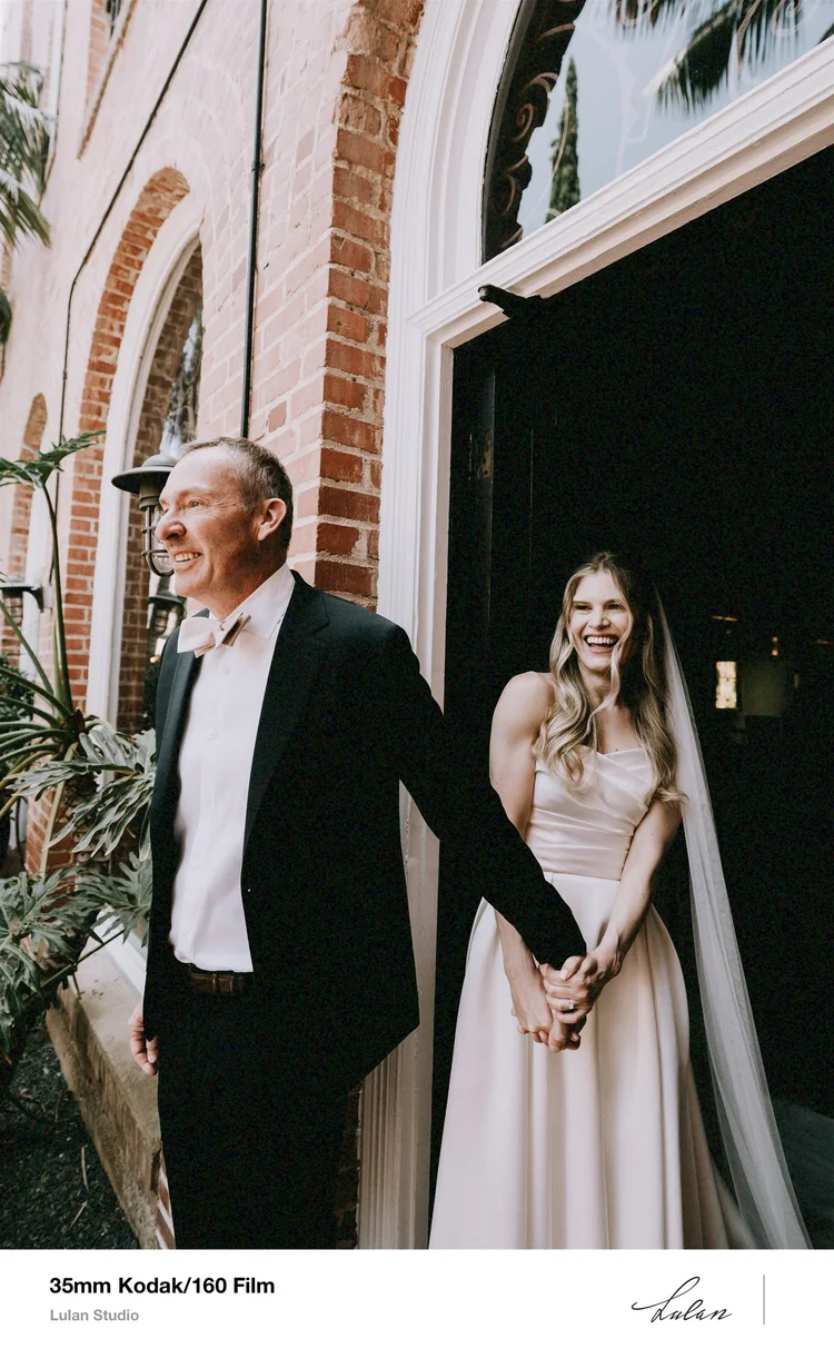 Film print: a bride and her father walking hand in hand along a brick building