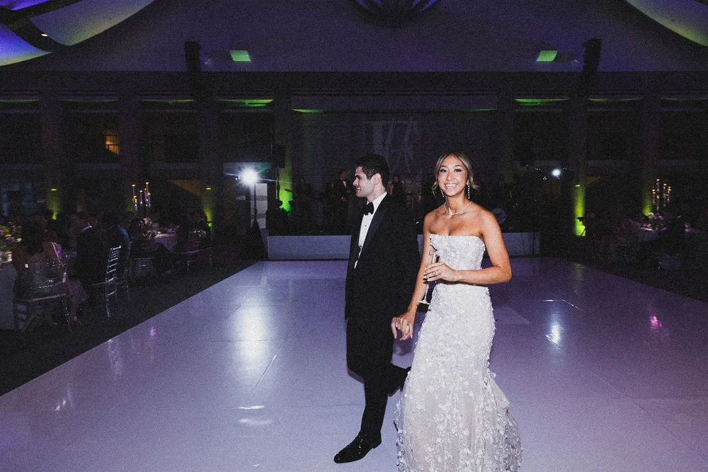 Couple walking across a white dance floor at their evening reception, the room washed in colored uplighting