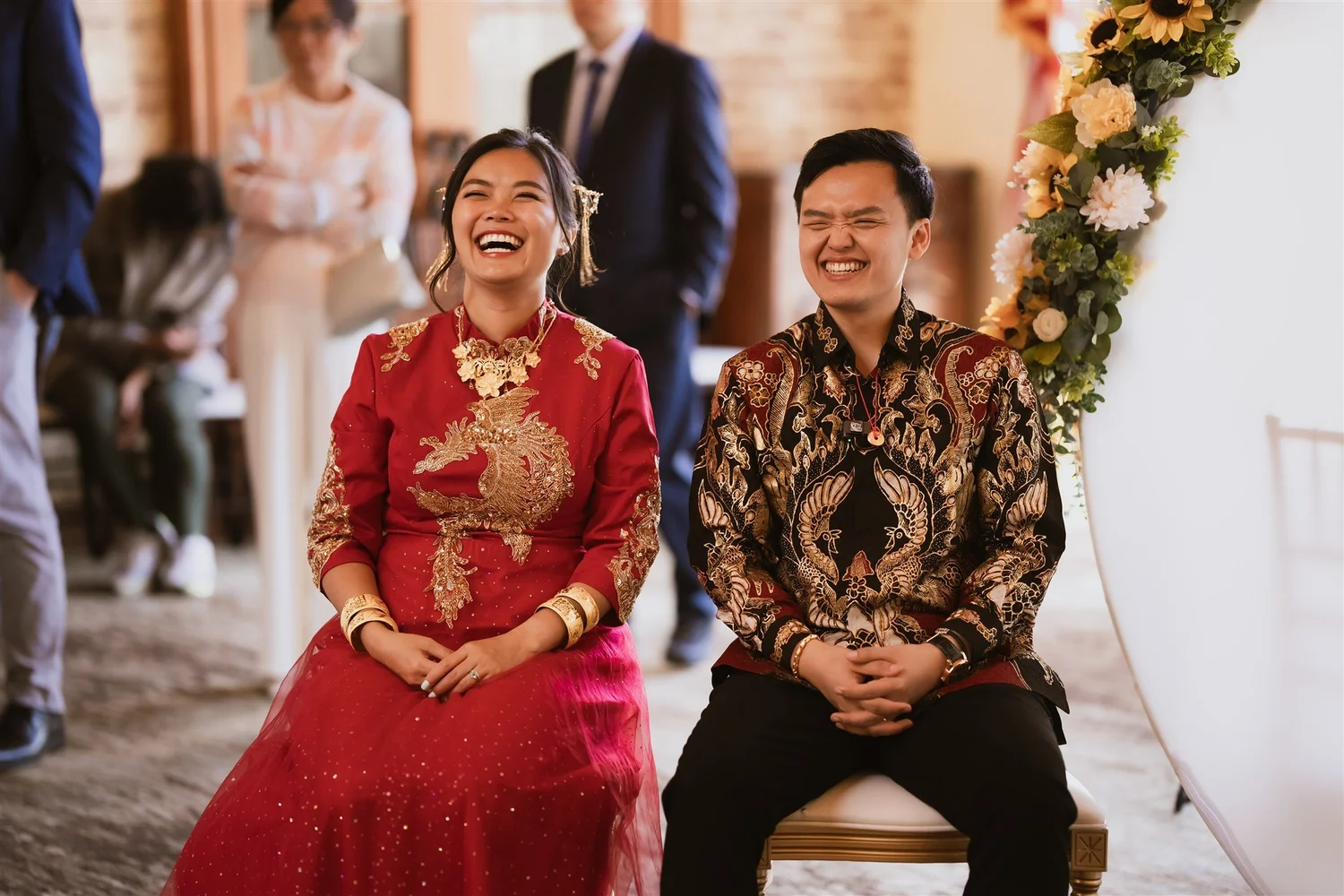 A couple in a red Chinese wedding dress and a batik shirt seated together during their tea ceremony in Los Angeles