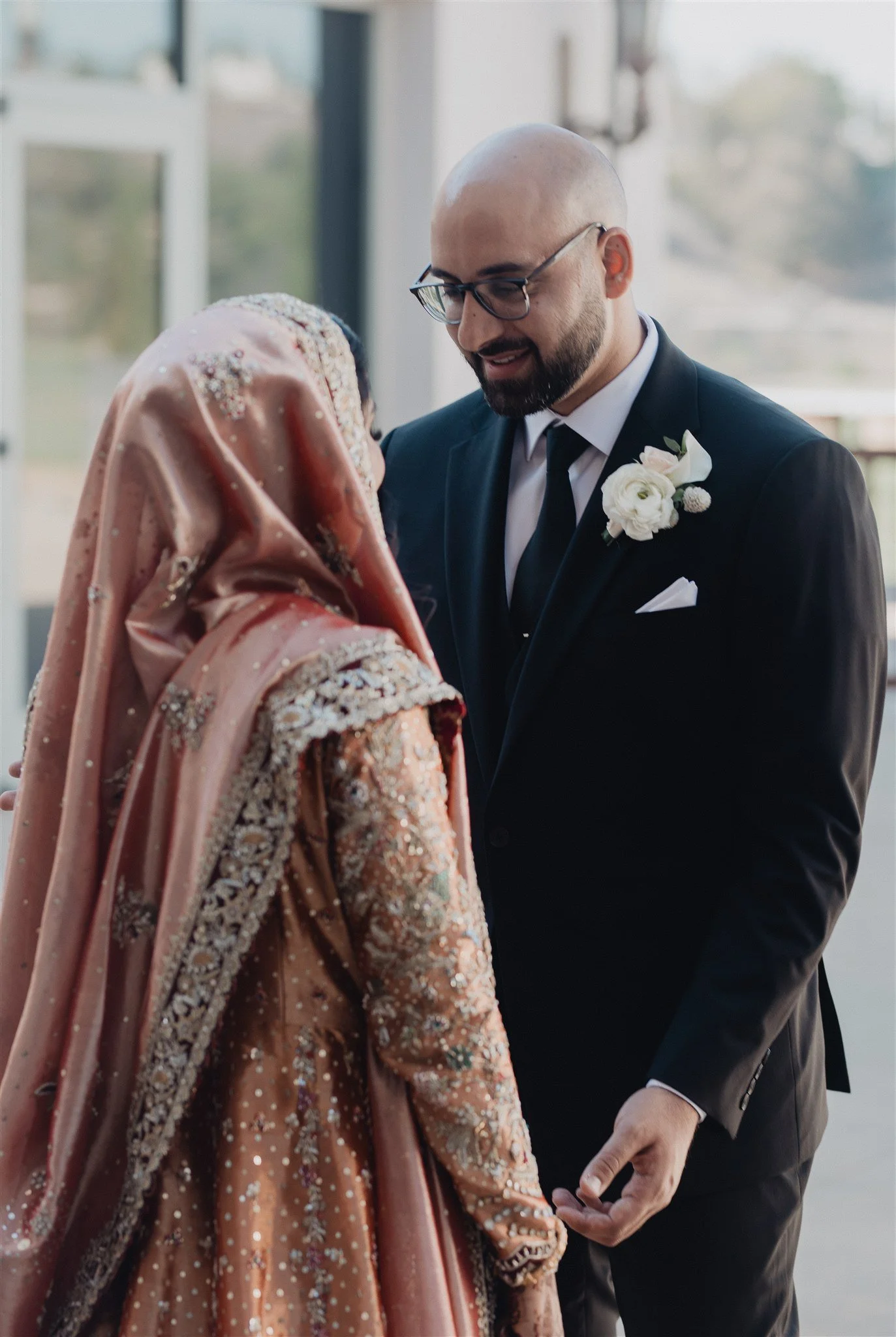 First look between a bride in a pink embroidered dupatta and her groom in a dark suit, beside a bright window