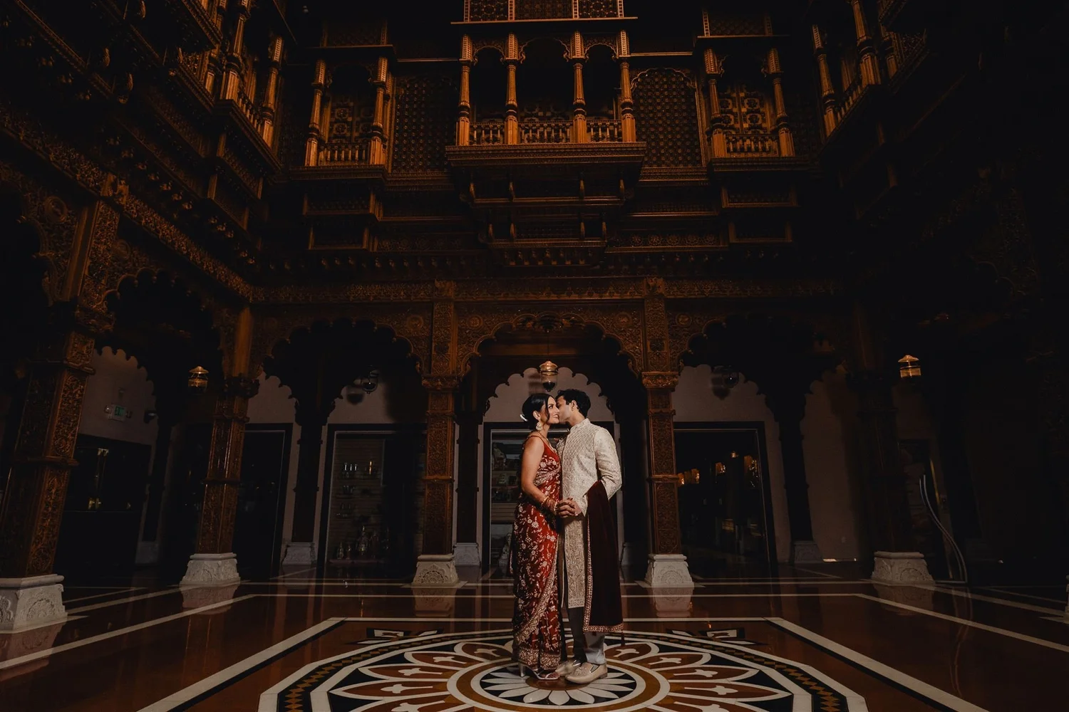 Bride and groom in traditional Indian wedding attire standing in an ornately carved hall