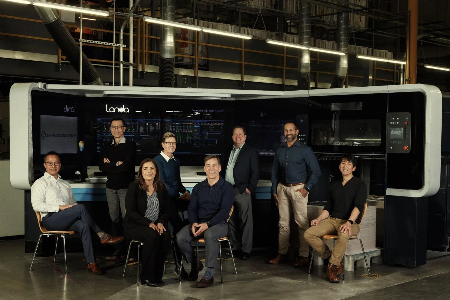 Group of nine professionals posing in front of a large, high-tech control panel or server machine in an industrial or research facility.