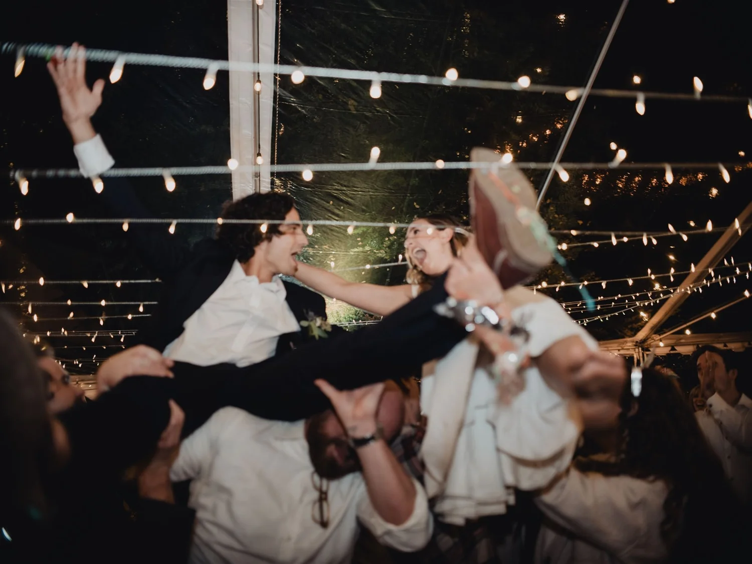 Wedding guests lifting the bride and groom into the air under string lights at a night-time reception