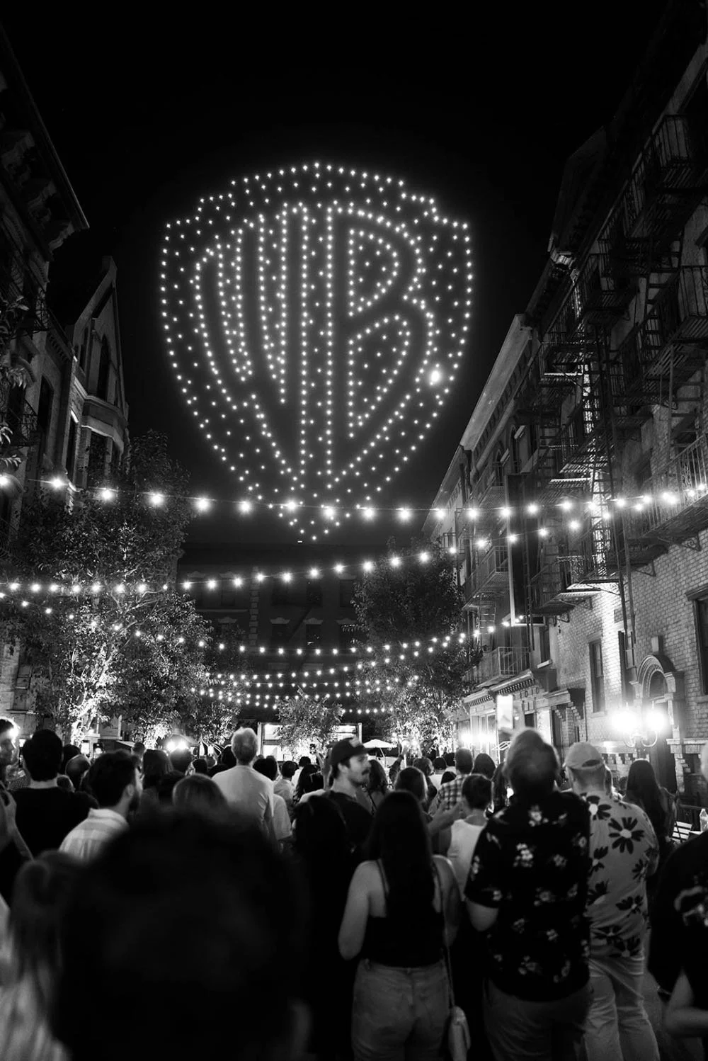 Nighttime street scene with many people gathered, decorative string lights hanging overhead, and a large illuminated Warner Bros. logo hot air balloon display in the sky.