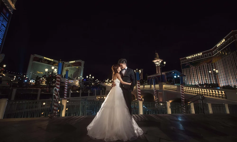 A bride and groom embrace and smile at night on a bridge, with the illuminated Mirage and Treasure Island hotels and casino in the background on the Las Vegas Strip.