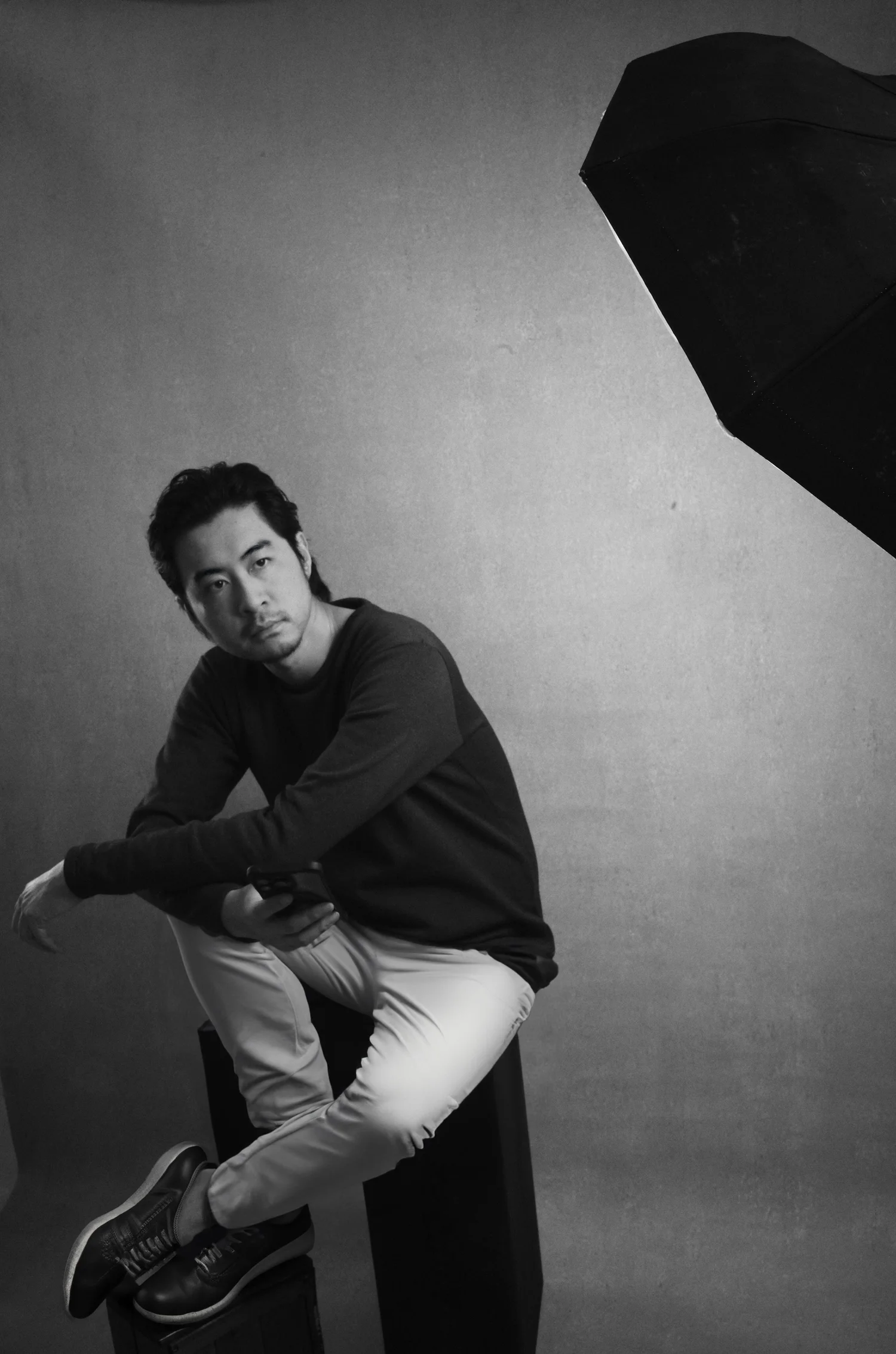 Black and white 35mm film portrait of a seated man in a dark sweater, lit against a plain studio backdrop
