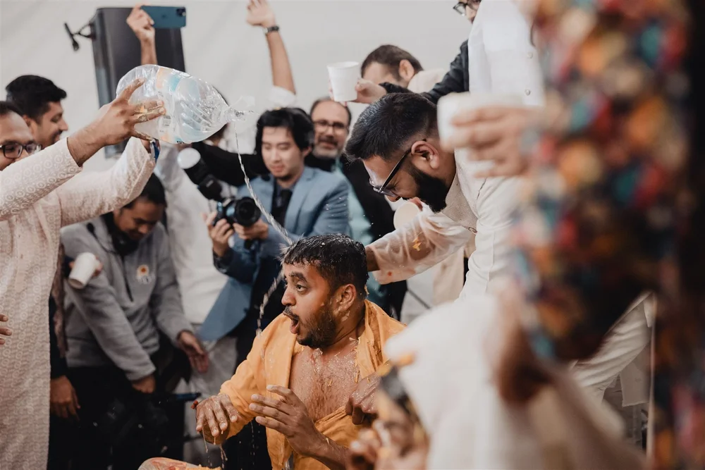 Friends pouring water over a laughing groom at a haldi celebration