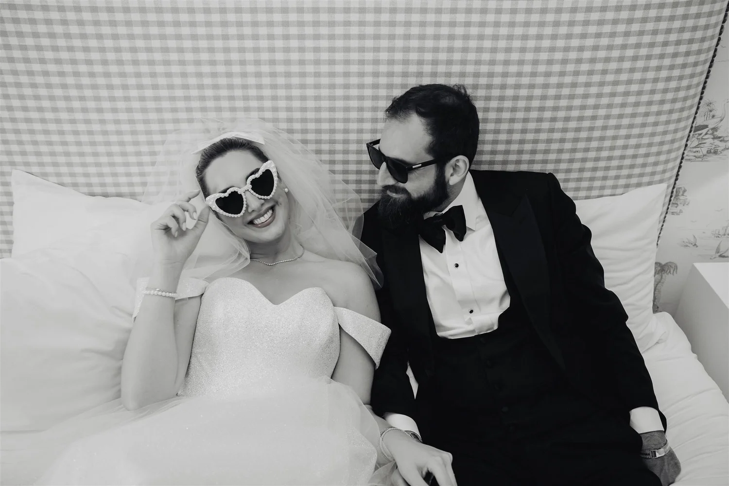 Black-and-white photo of a bride in heart-shaped sunglasses laughing beside her groom on a hotel bed