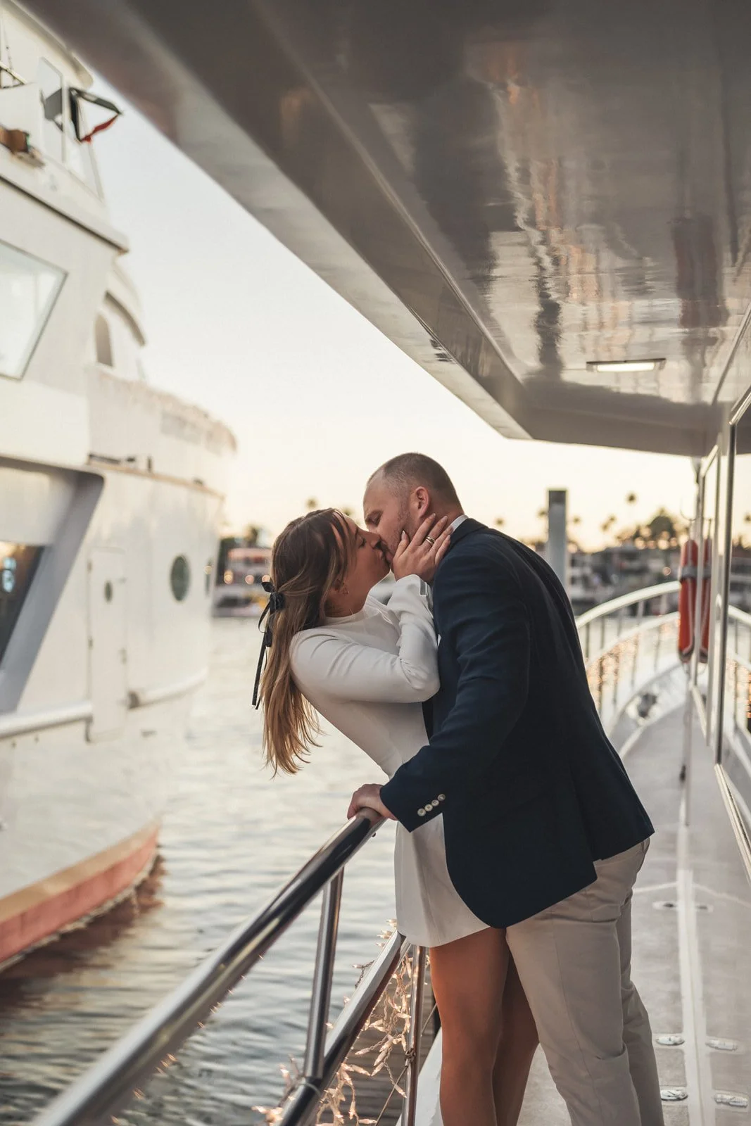 Couple kissing on the deck of a yacht at sunset in a Southern California marina, string lights along the rail