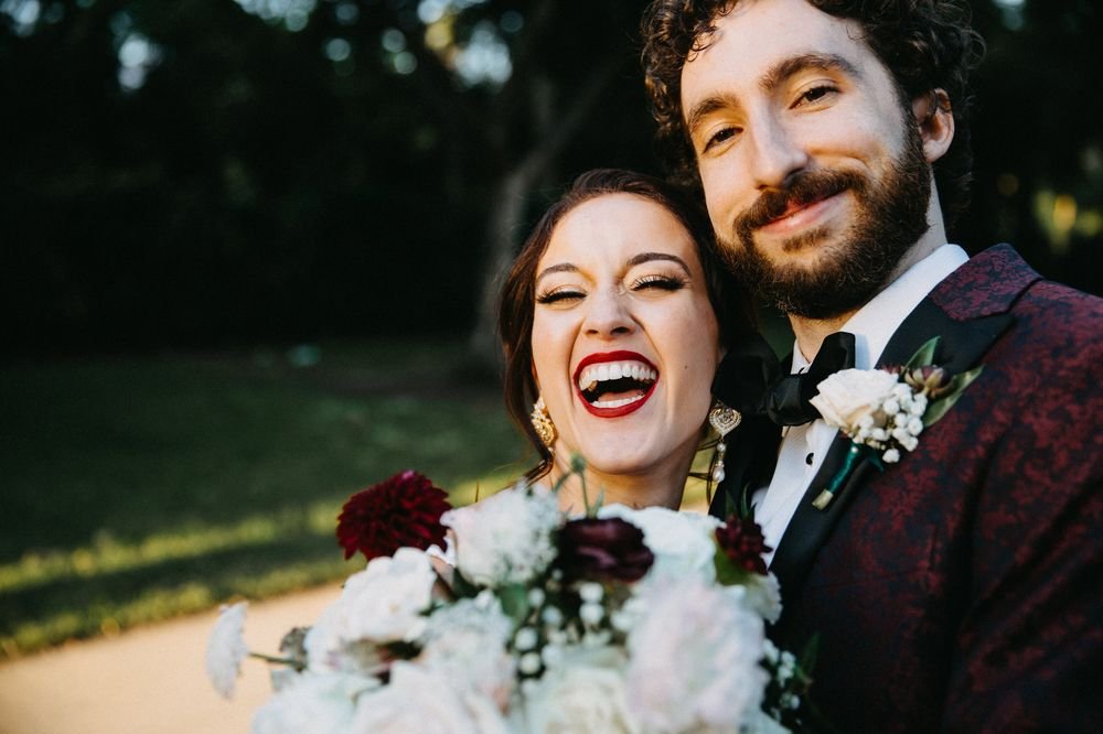 Laughing couple close to the camera, the bride holding a white and burgundy bouquet