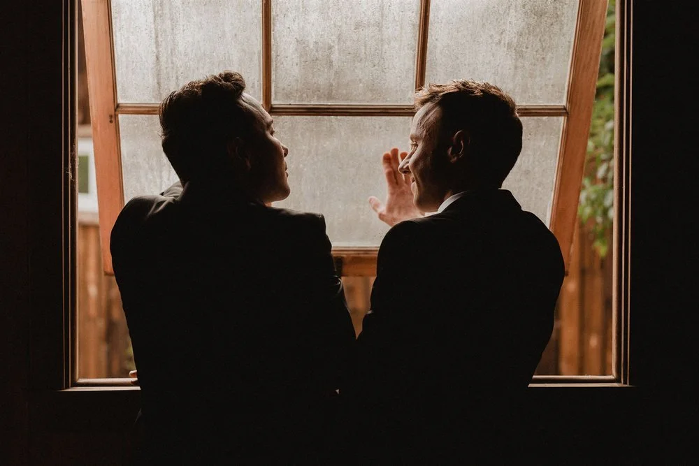 Two grooms in suits silhouetted against a large industrial window, talking to each other before their wedding ceremony