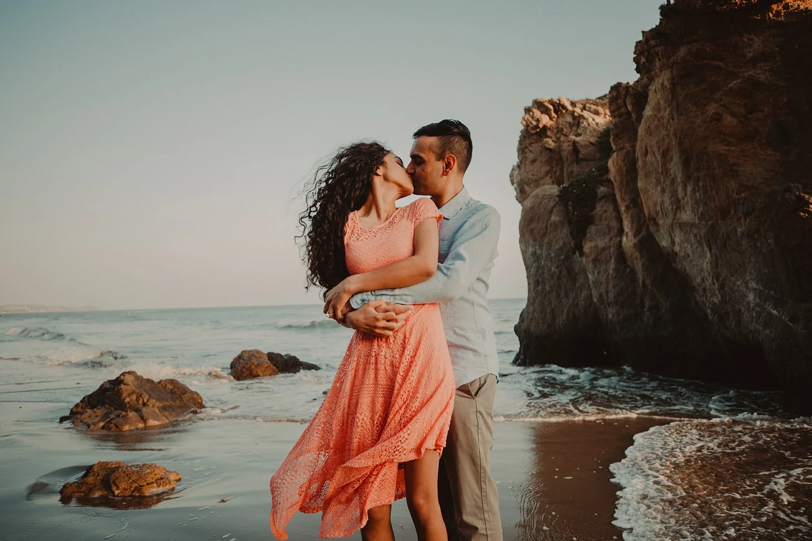 Couple in a coral dress and linen shirt kissing at the waterline below the sea arch at El Matador State Beach, Malibu