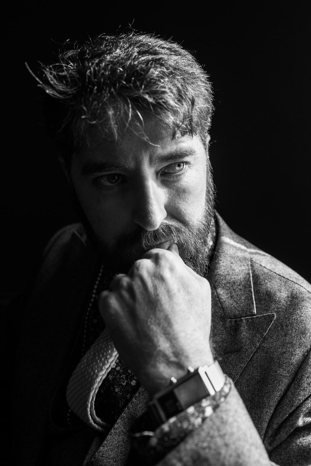 Black and white portrait of a bearded man resting his chin on his hand, lit by a single hard light