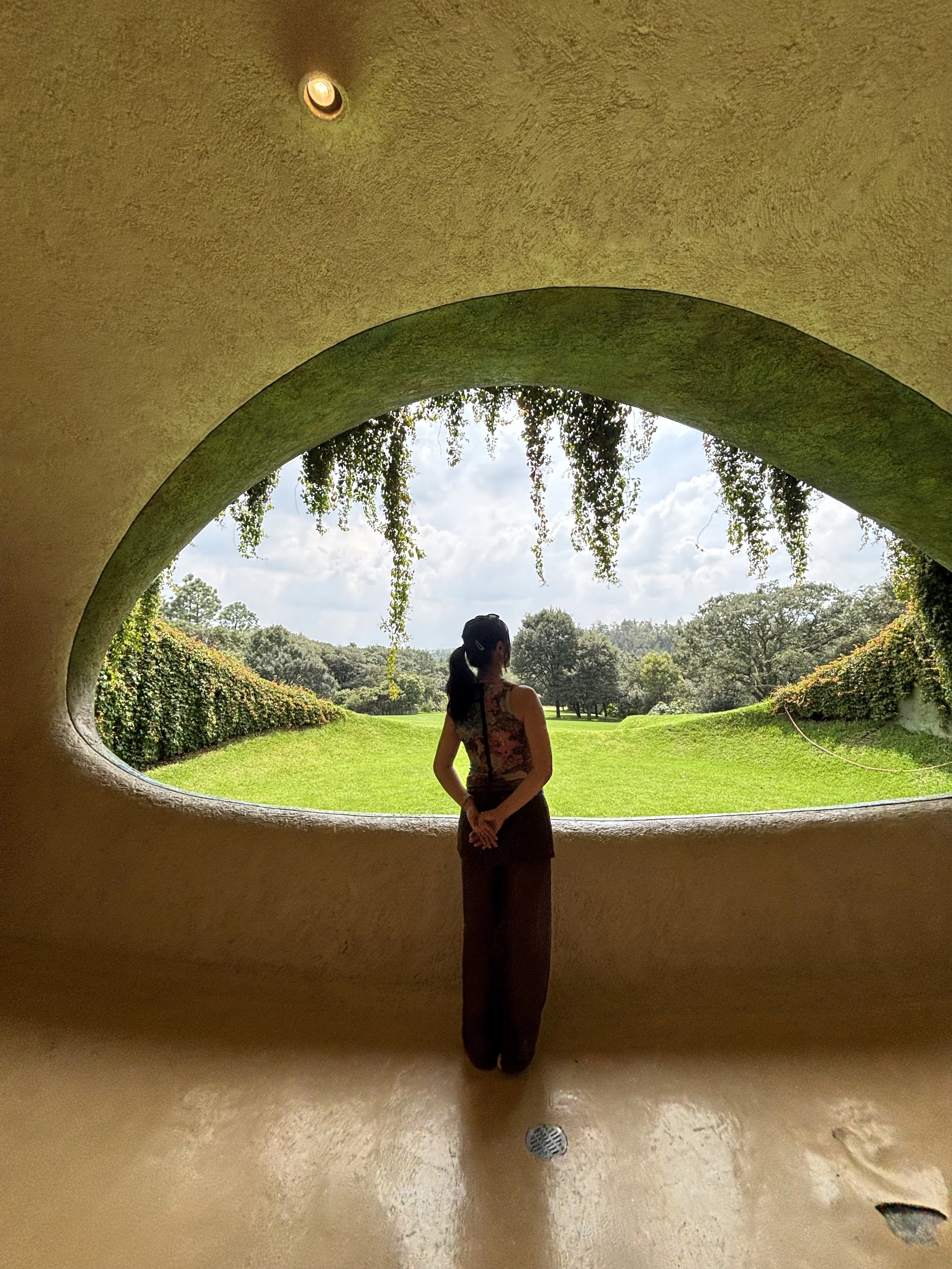 A person stands in a rounded earthen room, looking through an eye-shaped opening onto a green lawn and trees in Mexico City.