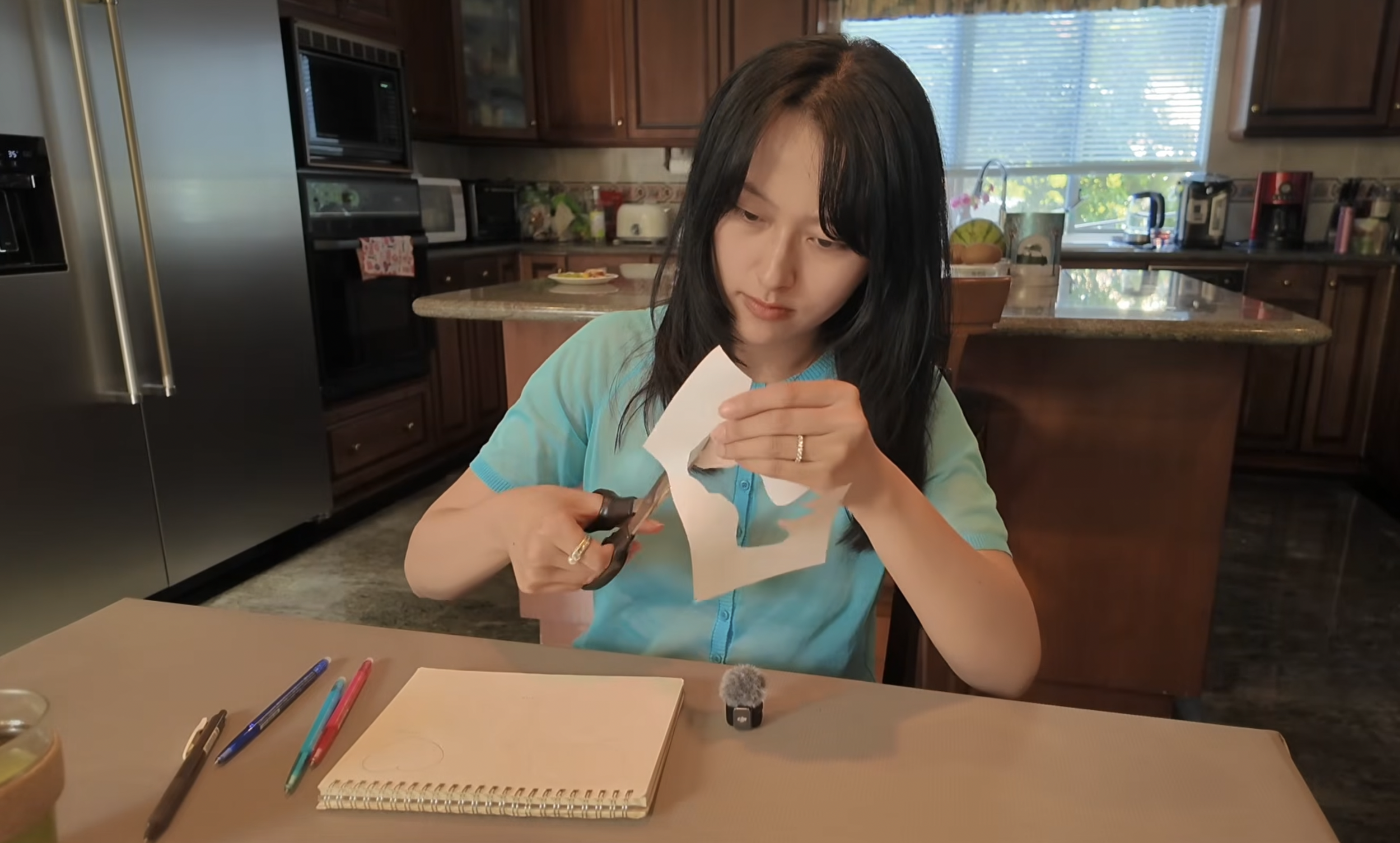 an asian female art therapist cutting a piece of paper and making art. she is wearing a bright turquoise blue shirt and there are colourful pens and matcha around her.