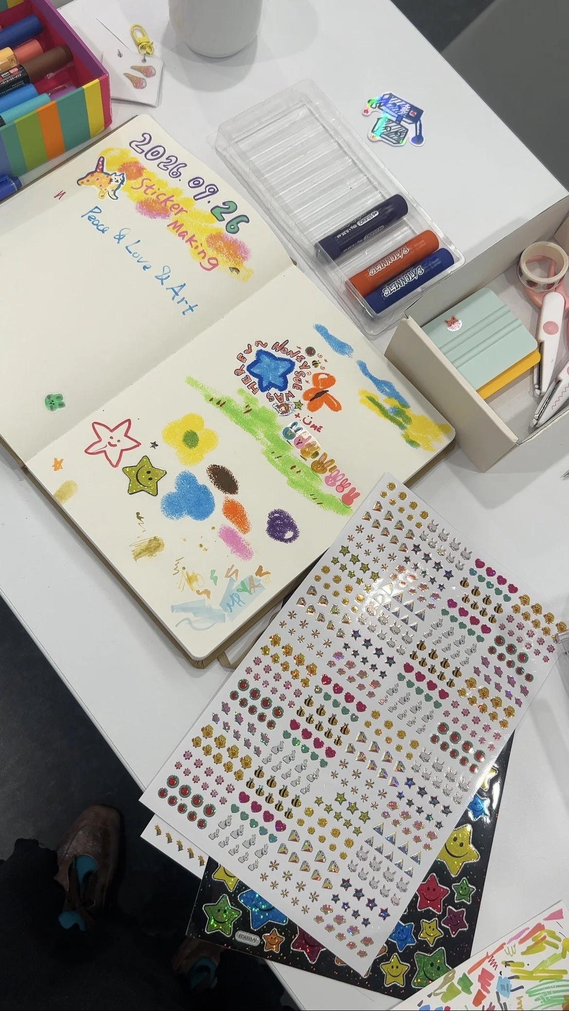 An open sketchbook on a white table at a sticker-making workshop, filled with colourful paint-stick drawings of stars, flowers and clouds, beside sheets of sparkly stickers, paint sticks and scissors.