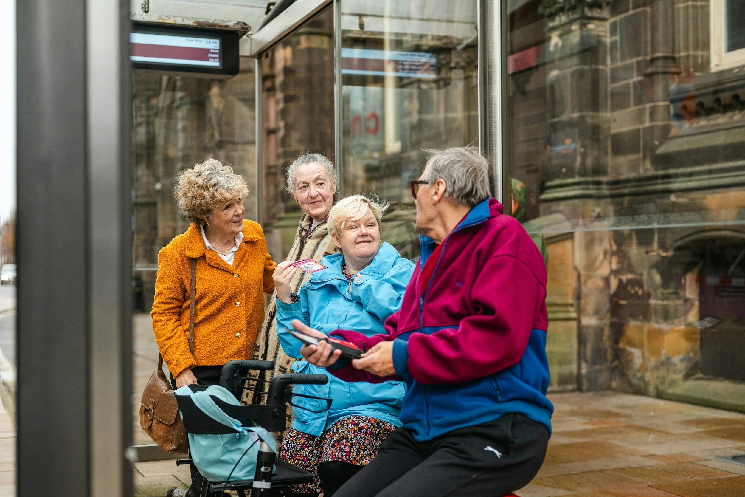 Three older ladies and an elderly man having a chat at a bus stop against a stone building, it's been raining