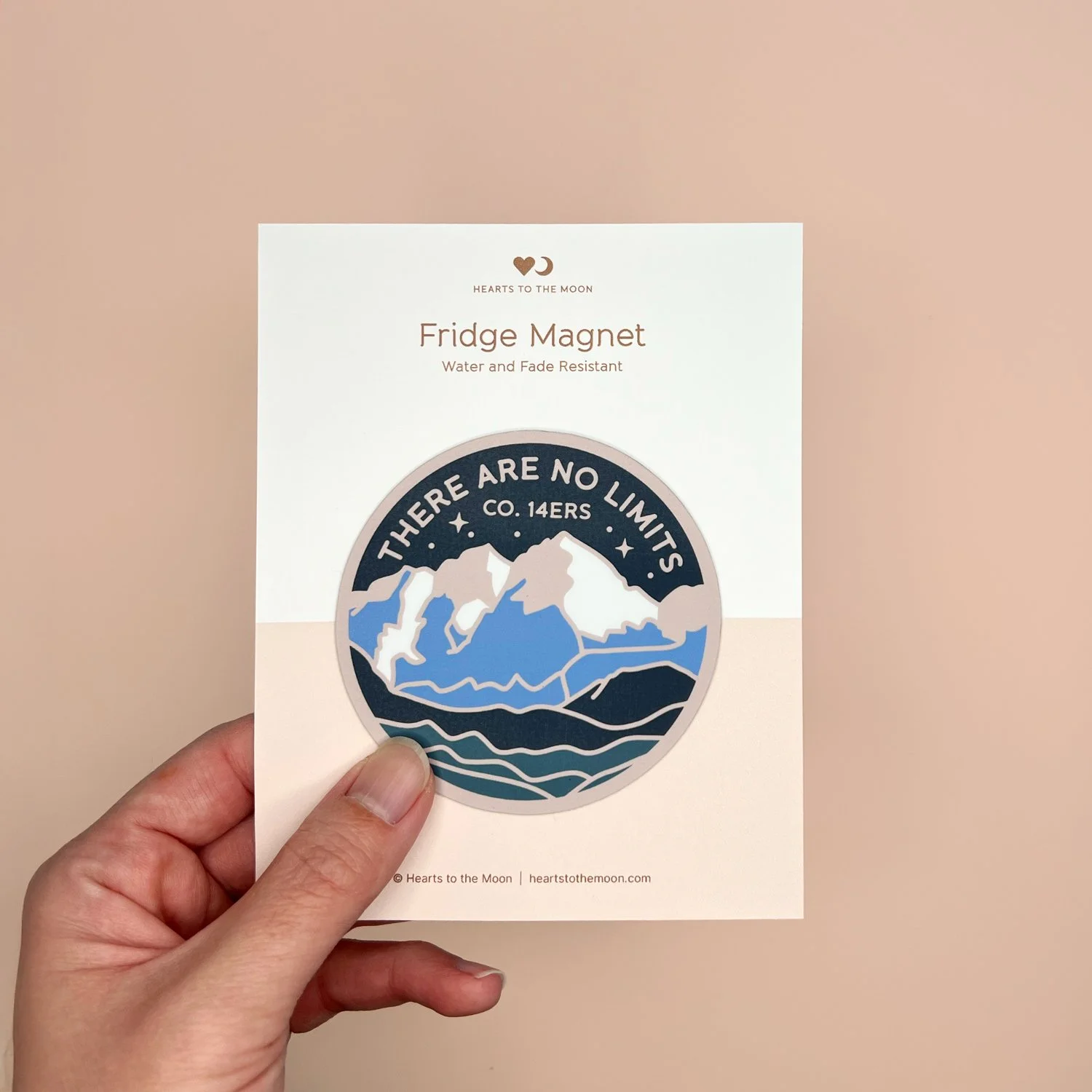 Hand holding a handmade 3-inch round mountain fridge magnet on backing card against a warm beige background.