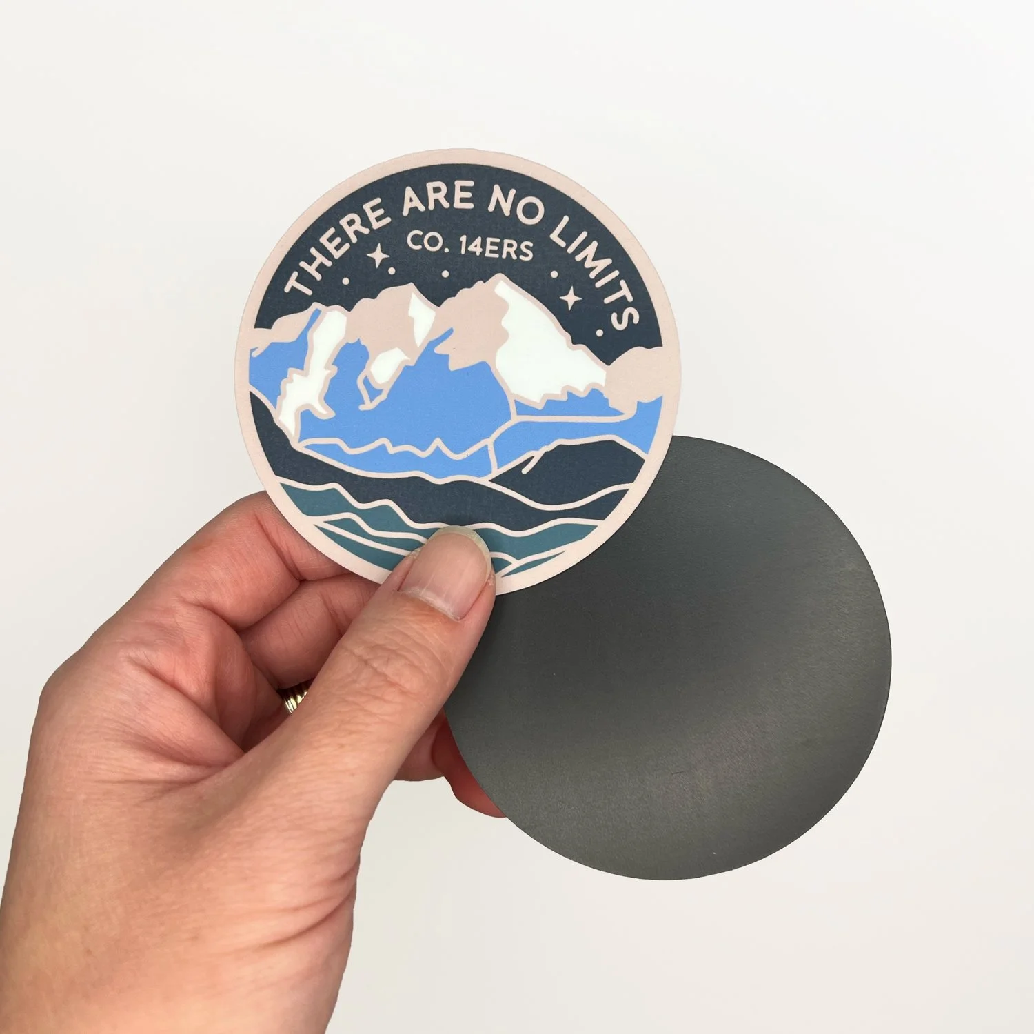 Hand holding a handmade 3-inch round mountain fridge magnet showing the illustrated front and plain magnetic back against a white background.