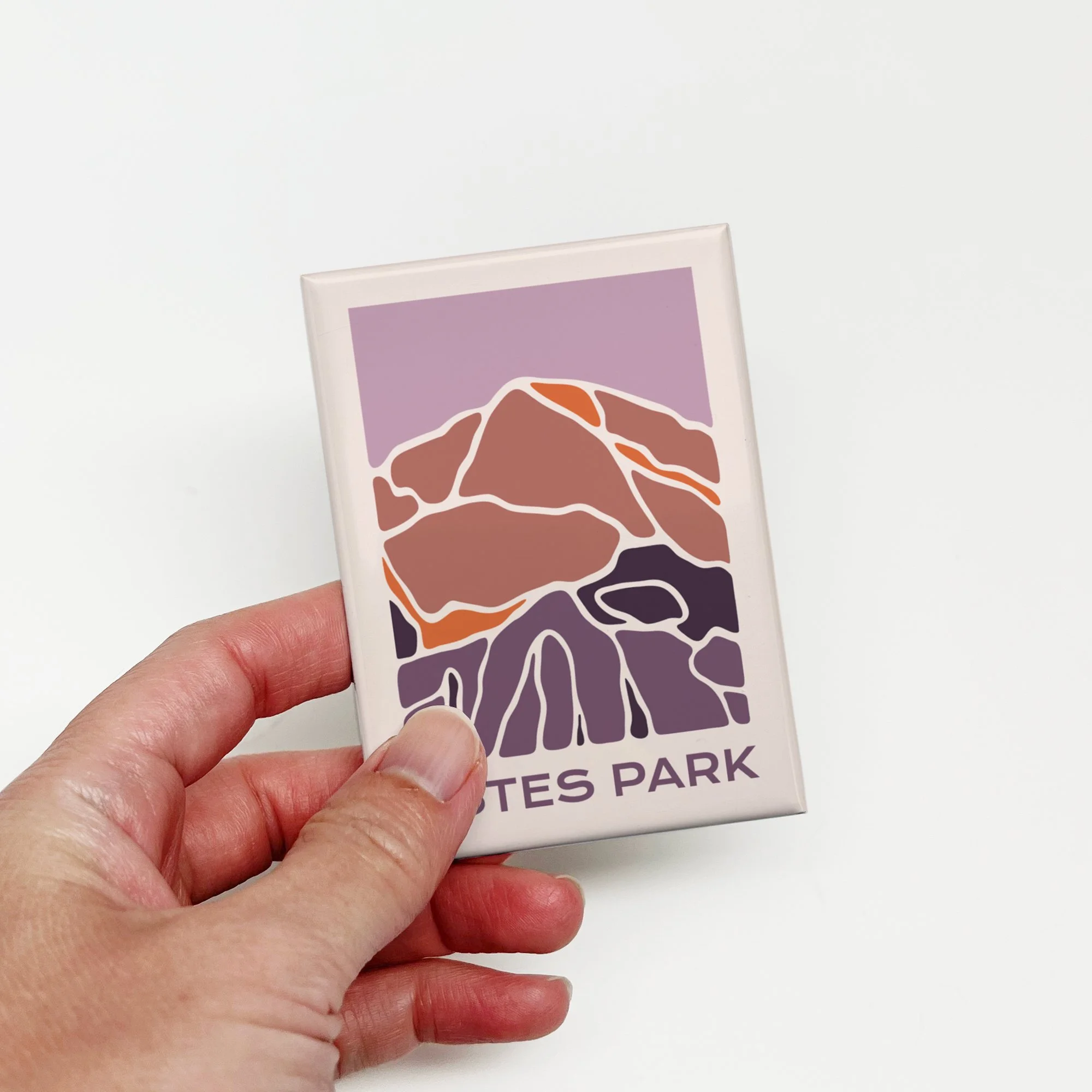 Hand holding a modern Estes Park magnet featuring an abstract mountain landscape in purple, mauve, and orange.