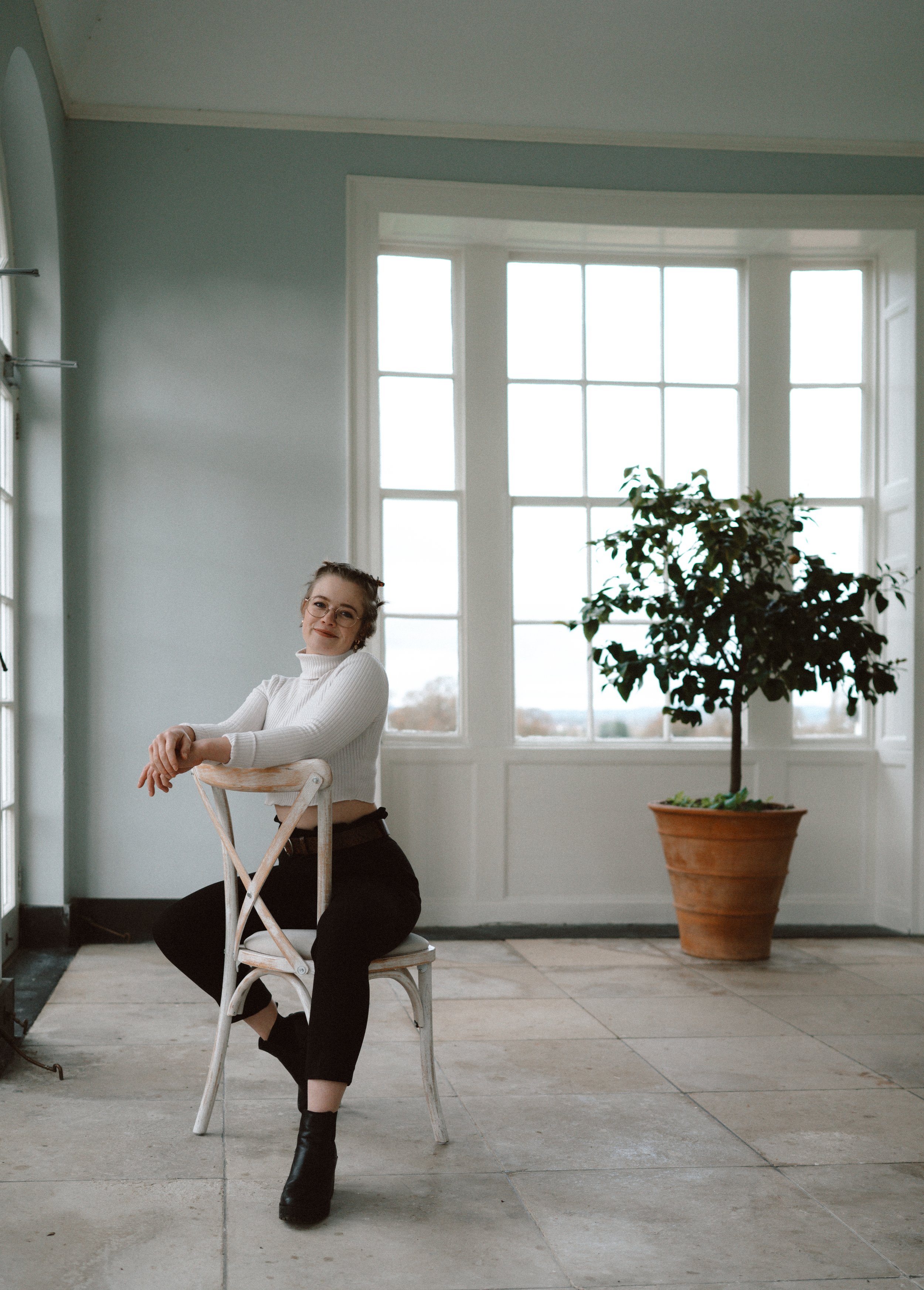 Photo of Alannah Veitch, a wedding content creator. She is a white woman in her twenties, wearing a white jumper and black trousers, sitting on a chair in a large empty room.