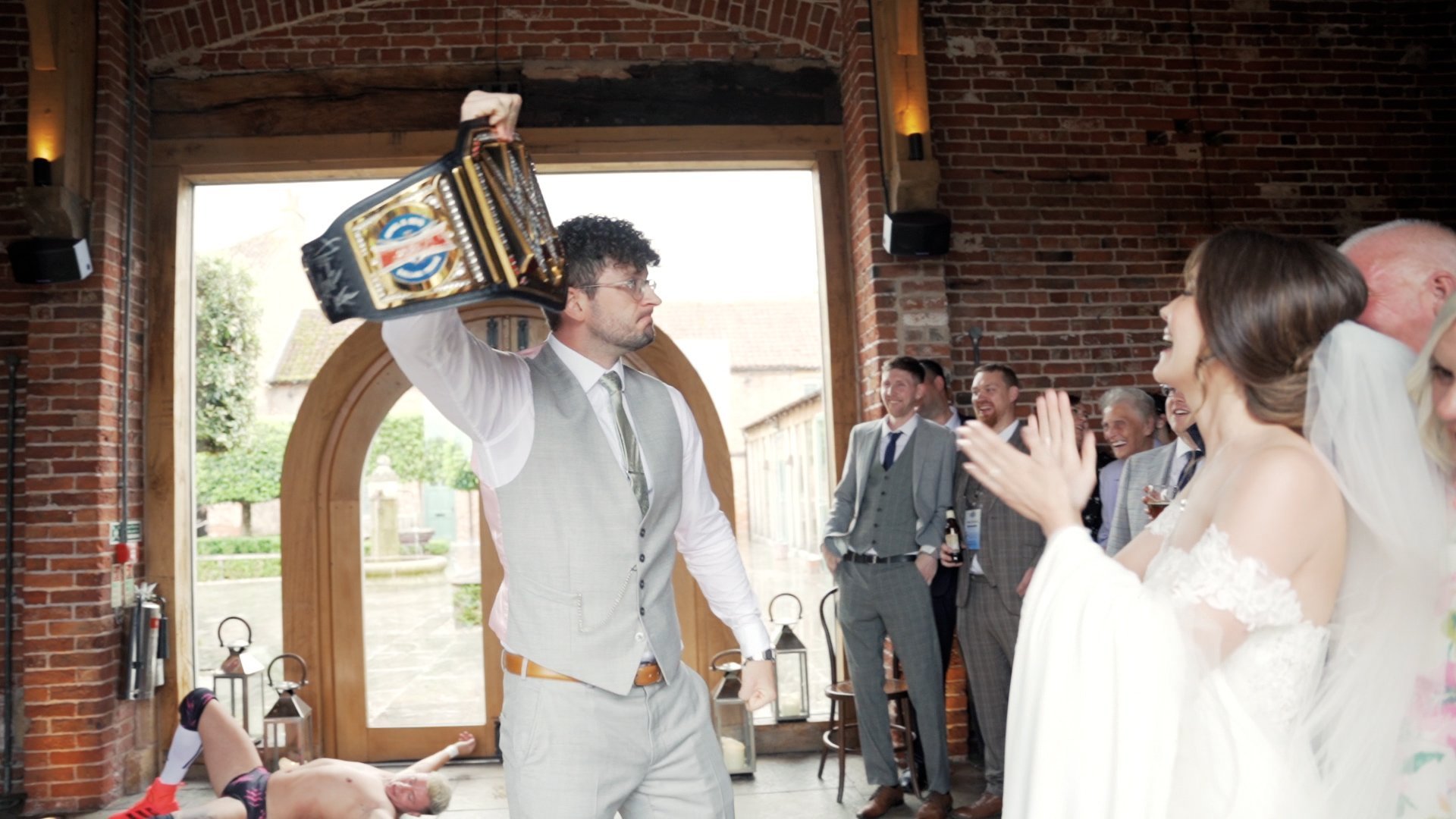 Groom at his wrestling themed wedding holding up a wrestling belt as wedding guests and bride cheer