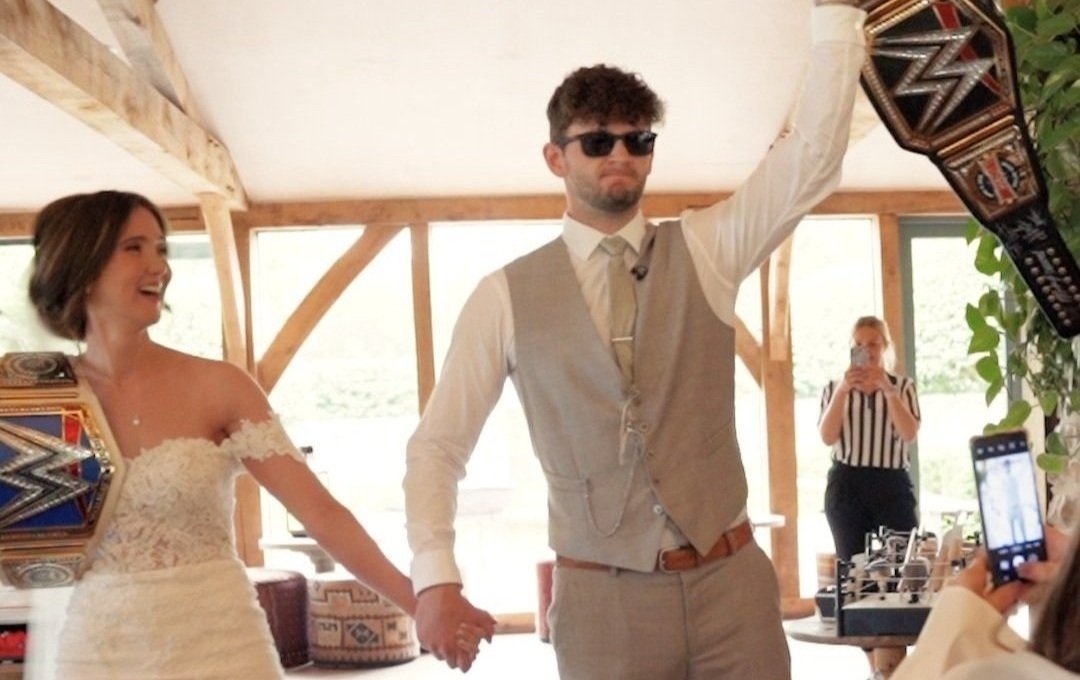 Bride and groom entering their WWE themed wedding reception holding wrestling belts up in the air. The groom is wearing dark sunlgasses and a grey suit and the couple are holding hands