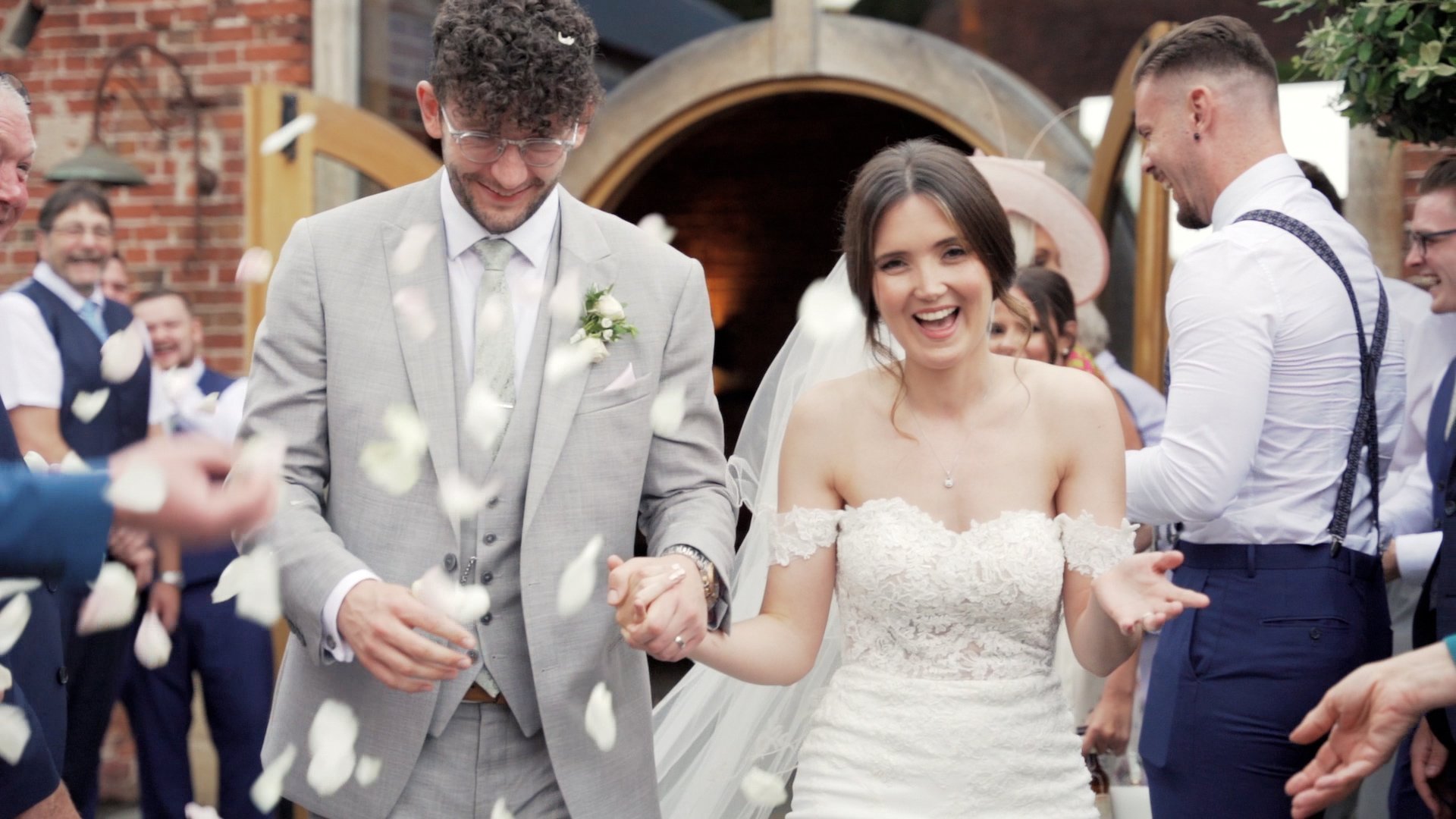 Bride and groom walking through guests throwing confetti. The groom is wearing a light grey 3-piece suit and the Bride is wearing a white lace wedding dress