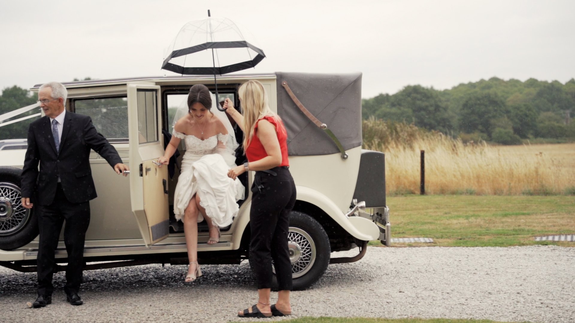 A beautiful bride stepping out of a wintage cream-coloured wedding car while a wedding guest holds an umbrella over her head