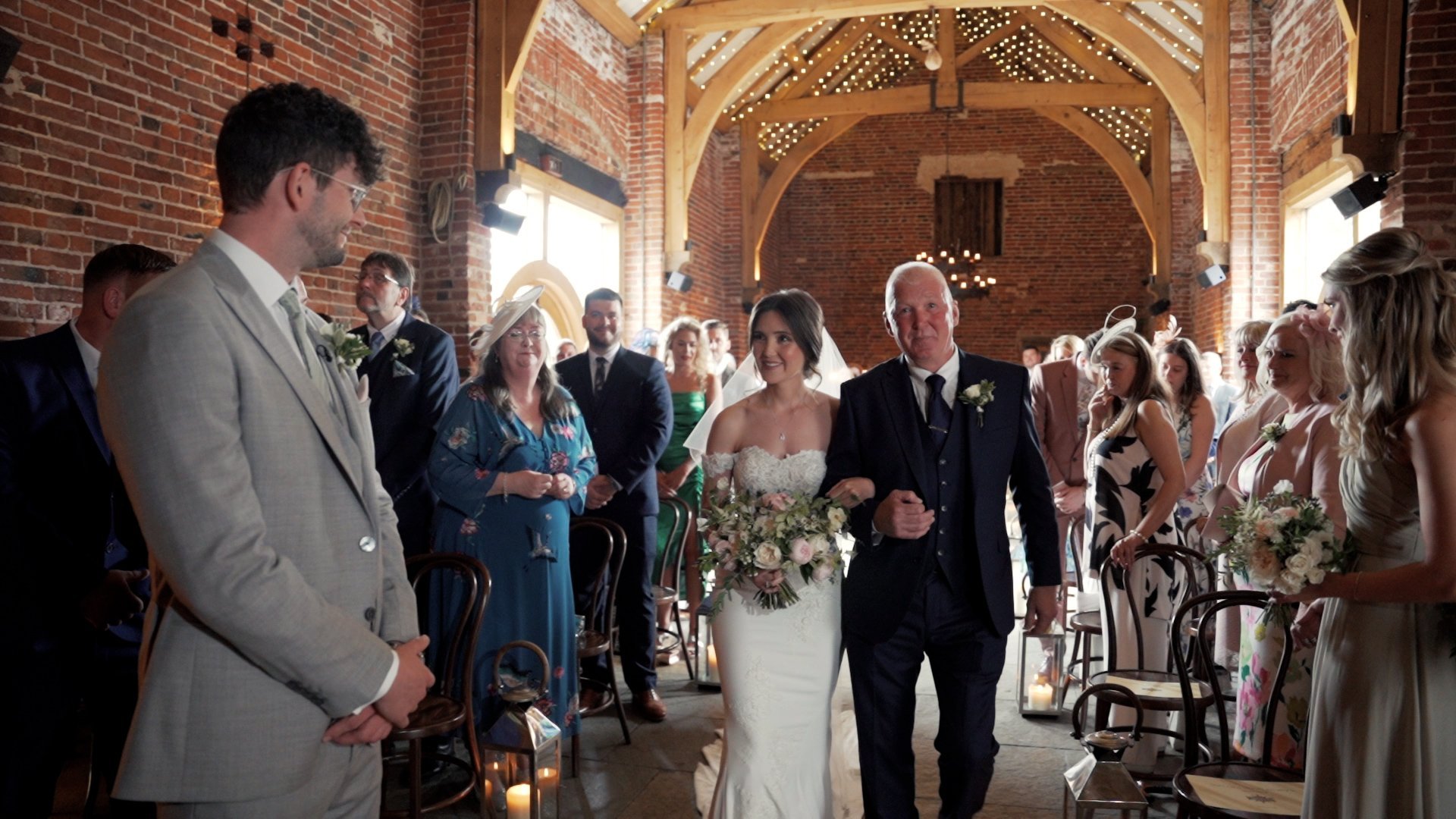 Bride is being walked down the aisle by her Father inside a beautiful wedding venue. She is walking towards her Groom who looks on with a smile.