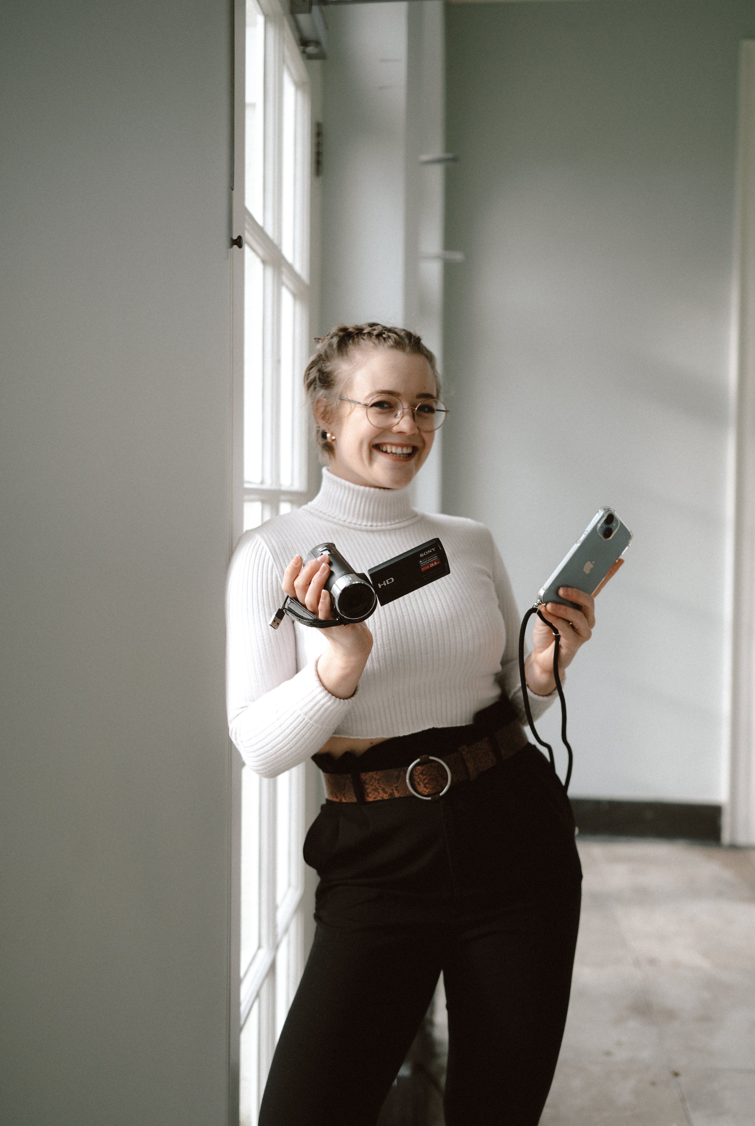 Photo of Alannah Veitch, a wedding content creator. She is a white woman in her twenties, wearing a white jumper and blac trousers. She is leaning against a door holding a video camera