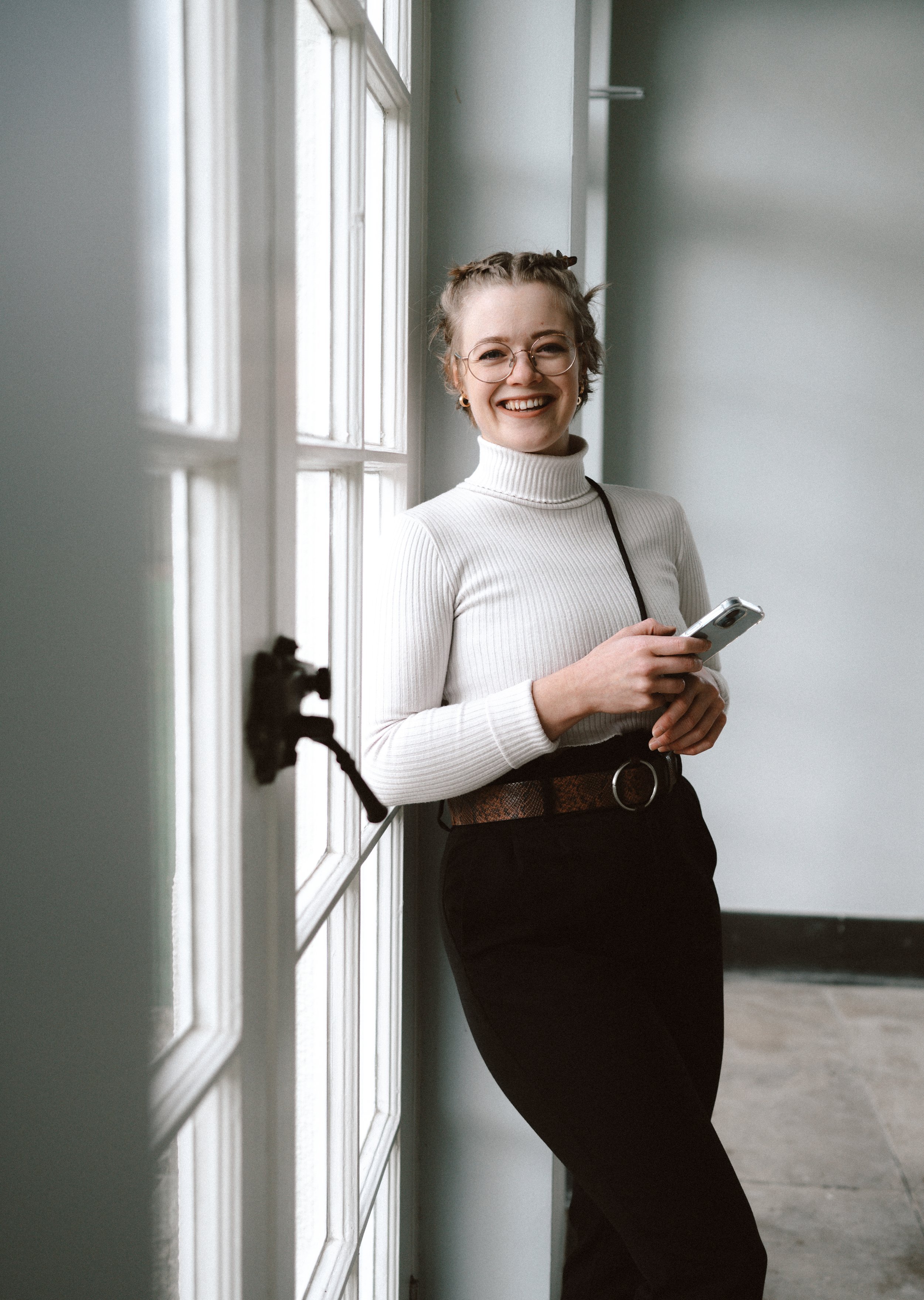 Photo of Allanah Veitch, a wedding content creator. She is a white woman in her twenties wearing a white jumper and black trousers. She is standing, leaning against a white door holding a smartphone