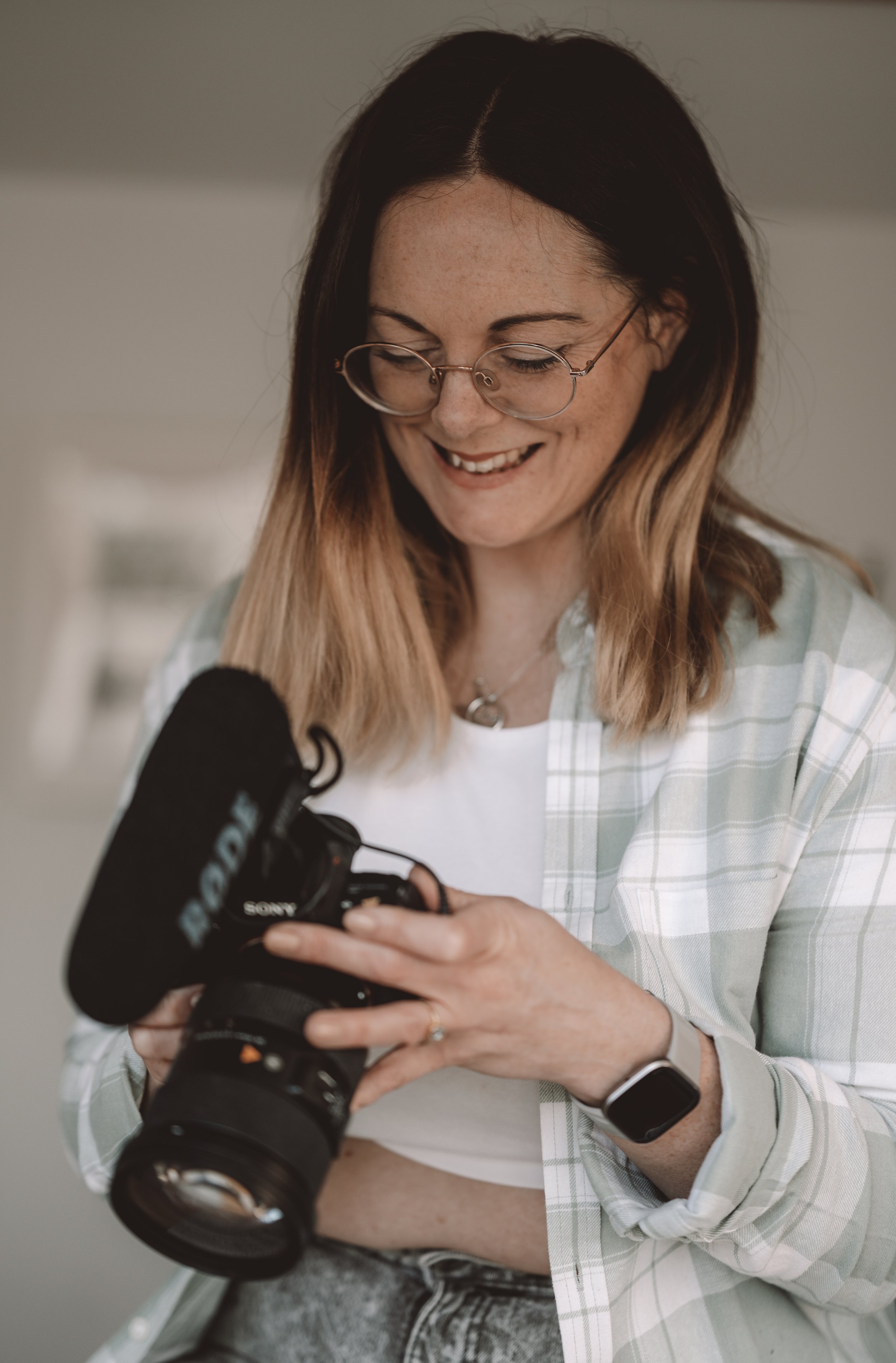Photo of Sally Johns, a wedidng videography. She is a young woman with long brown hair wearing casual clothes. She is looking at the back of a video camera while smiling.
