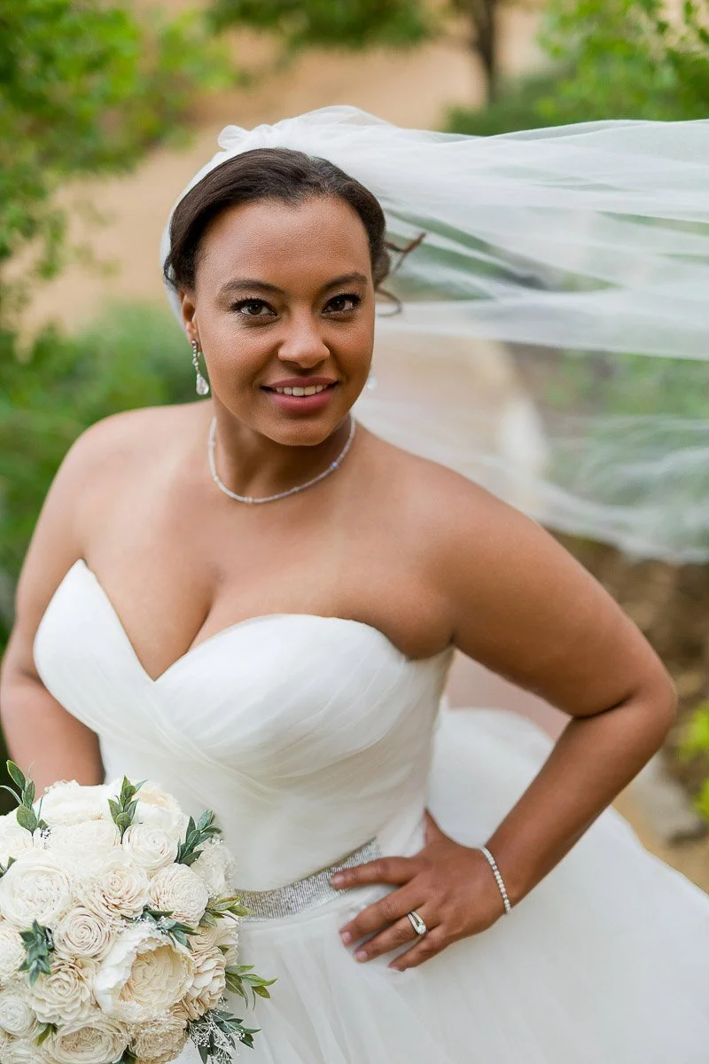 Bride smiling outdoors, wearing a strapless white gown with a flowing veil, holding a bouquet of white roses. Her hair is styled elegantly, and she exudes joy.