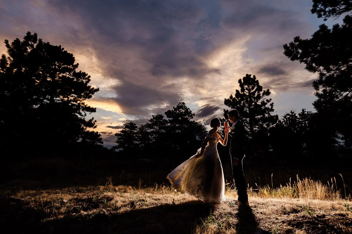 Silhouetted bride and groom share a moment at sunset, surrounded by trees. The dramatic sky and warm tones create a romantic, tranquil atmosphere.