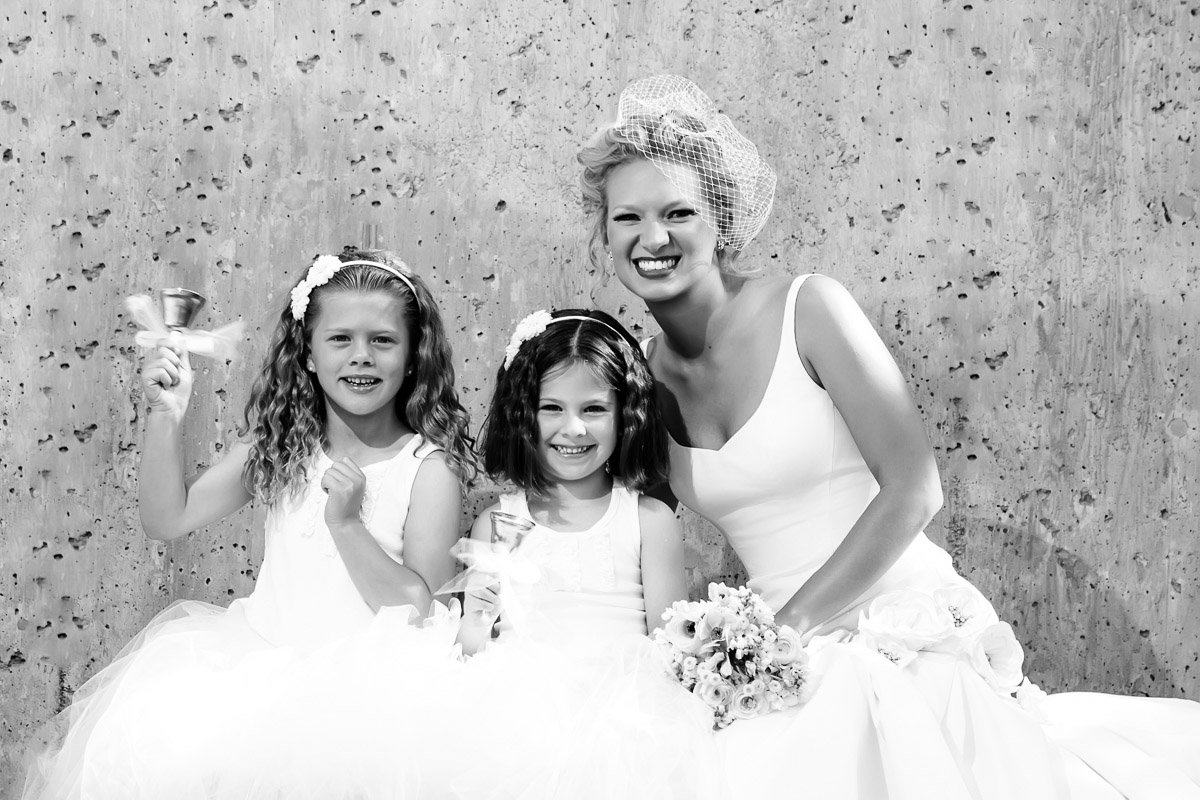 A bride in a white dress with a birdcage veil smiles alongside two young flower girls holding bells. All are joyful, set against a textured wall.