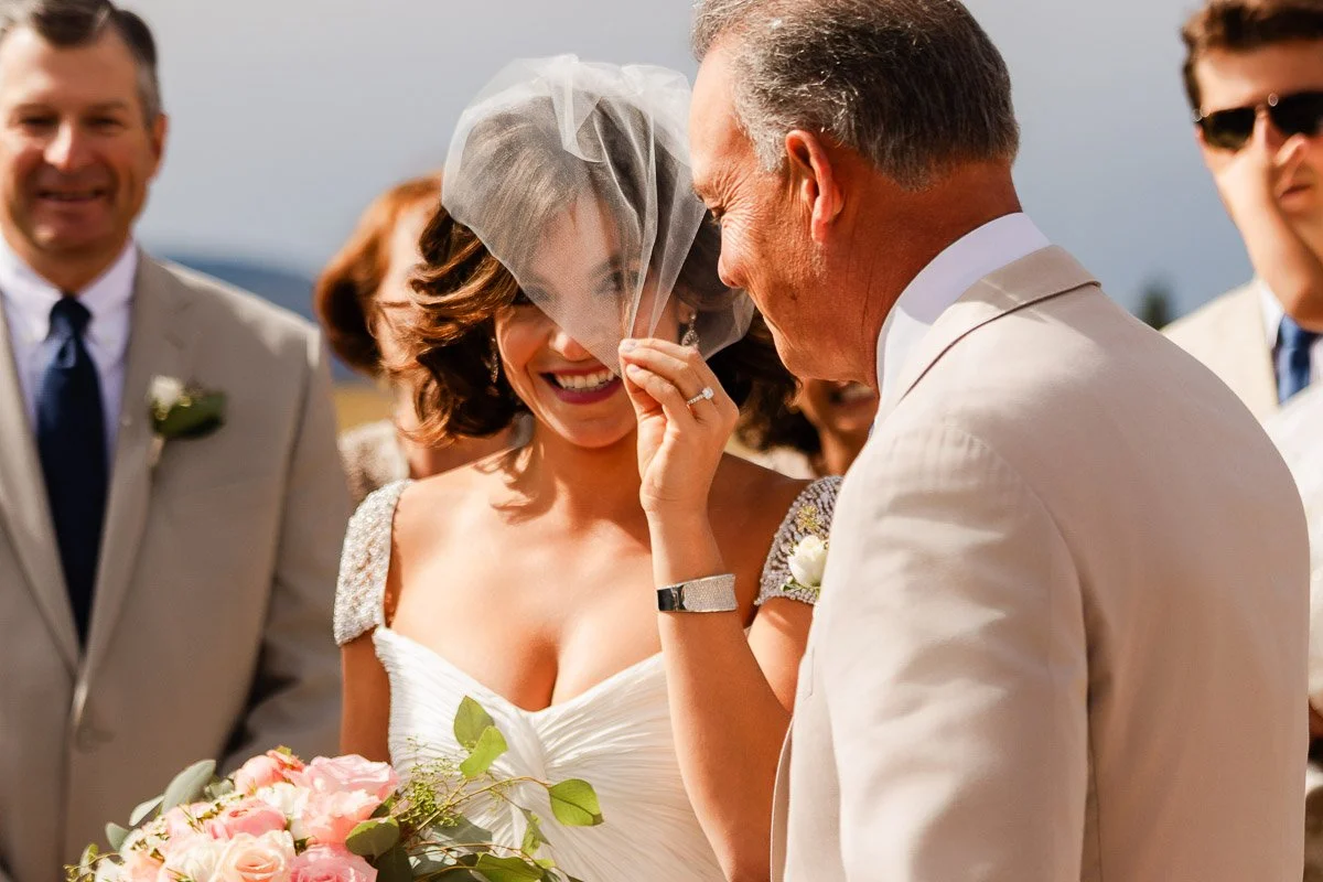 A joyful bride with a veil, holding a bouquet of pink roses, smiles at a man beside her in a light suit, surrounded by smiling guests.