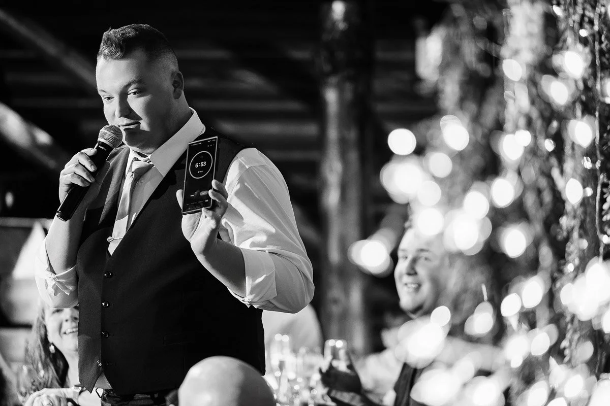 Man in a vest holds a microphone and phone timer showing 6:53 minutes, speaking at an event. Fairy lights blur in the foreground, guests smiling in the background. Black and white.